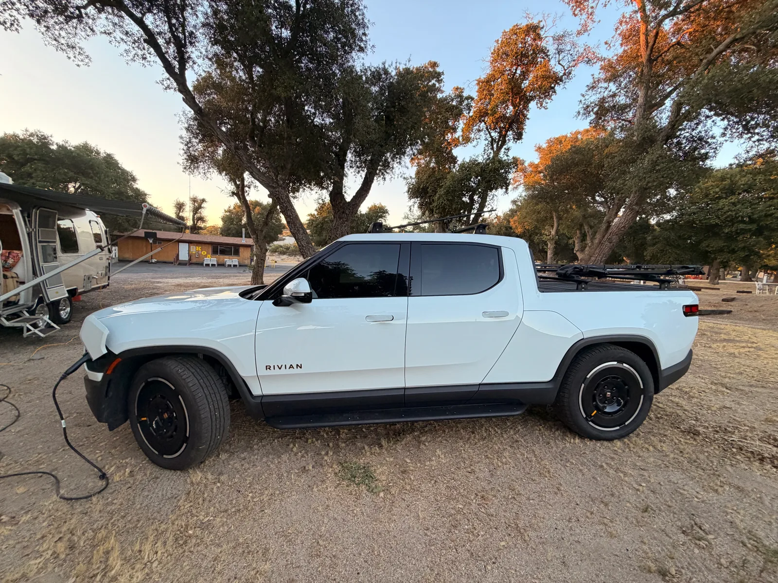 The white Rivian R1T plugged in at the Boulevard KOA campsite at golden hour, parked under California oaks with the e-bikes on the bed rack