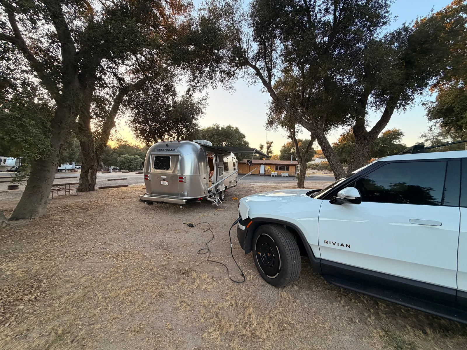 The 2024 Airstream International parked next to the white Rivian R1T at the Boulevard KOA campsite, both under oaks at sunset