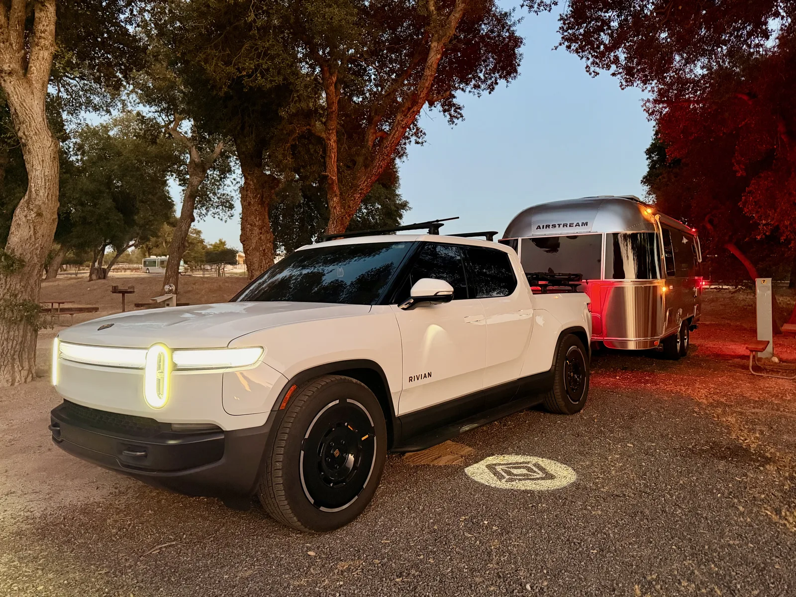 The white Rivian R1T with headlights on, hitched to the silver 2024 Airstream International at the Boulevard KOA campsite under California oaks at golden hour, ready to roll east