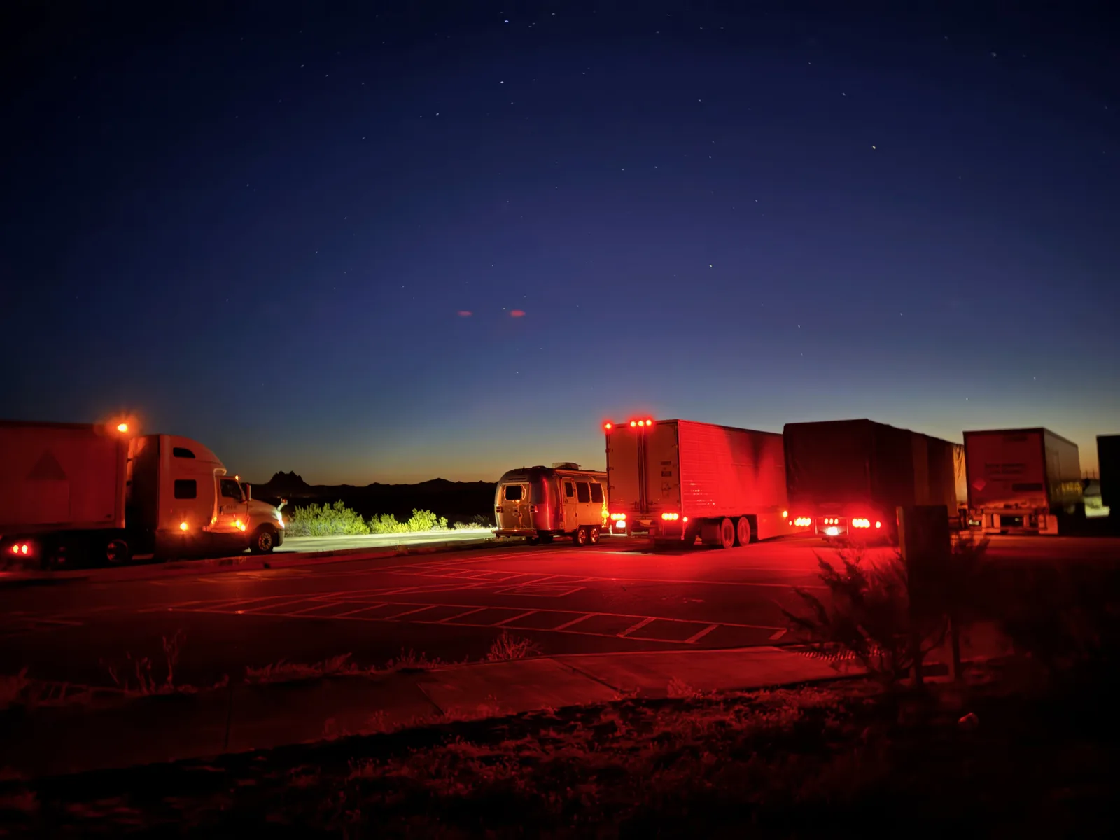 The silver Airstream International parked between two semi-truck trailers at an I-8 rest area east of Yuma at 1 AM, red running lights glowing in the desert dark with the Milky Way faintly overhead
