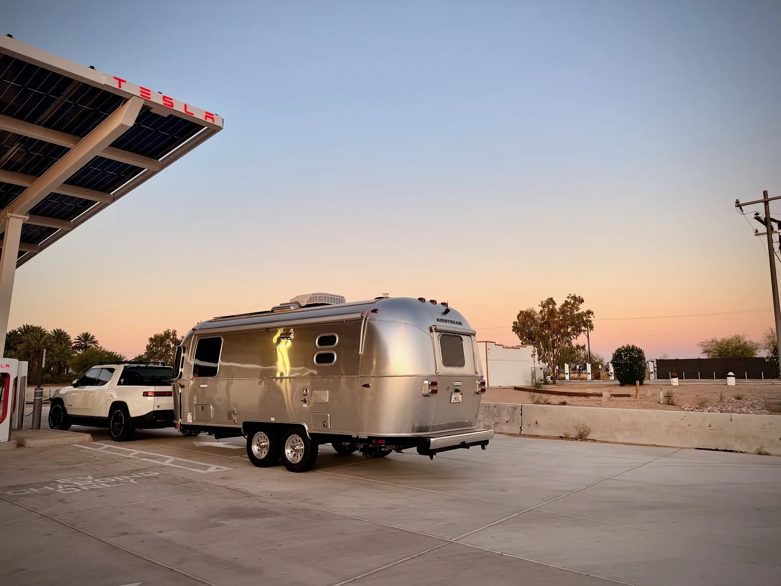 The silver 2024 Airstream International rear with SMUGL California vanity plate, parked alongside a TESLA solar-canopy Supercharger plaza at dawn, dozens of V4 pedestals lined up under the canopy