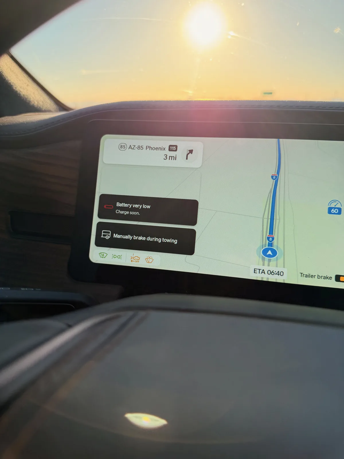 The Rivian R1T center display through the windshield at sunrise, navigation showing AZ-85 Phoenix exit 3 miles ahead, ETA 06:40, a "Battery very low" warning and a "Manually brake during towing" advisory