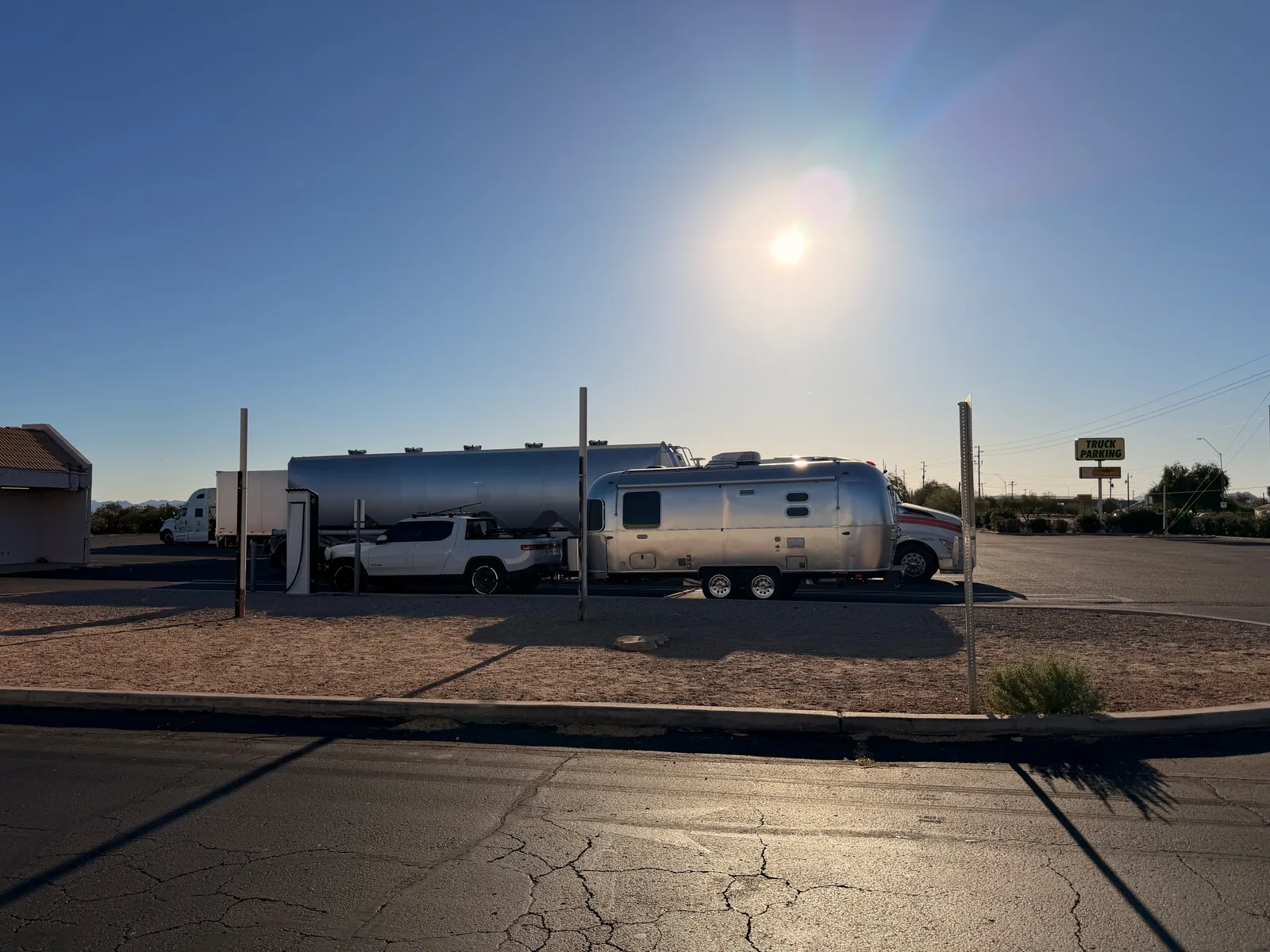 Wide view of the white Rivian R1T and silver Airstream International parked at the Gila Bend Rivian Adventure Network charger, with a tanker truck and Love's travel-plaza signage in the distance under a flare-lit blue sky