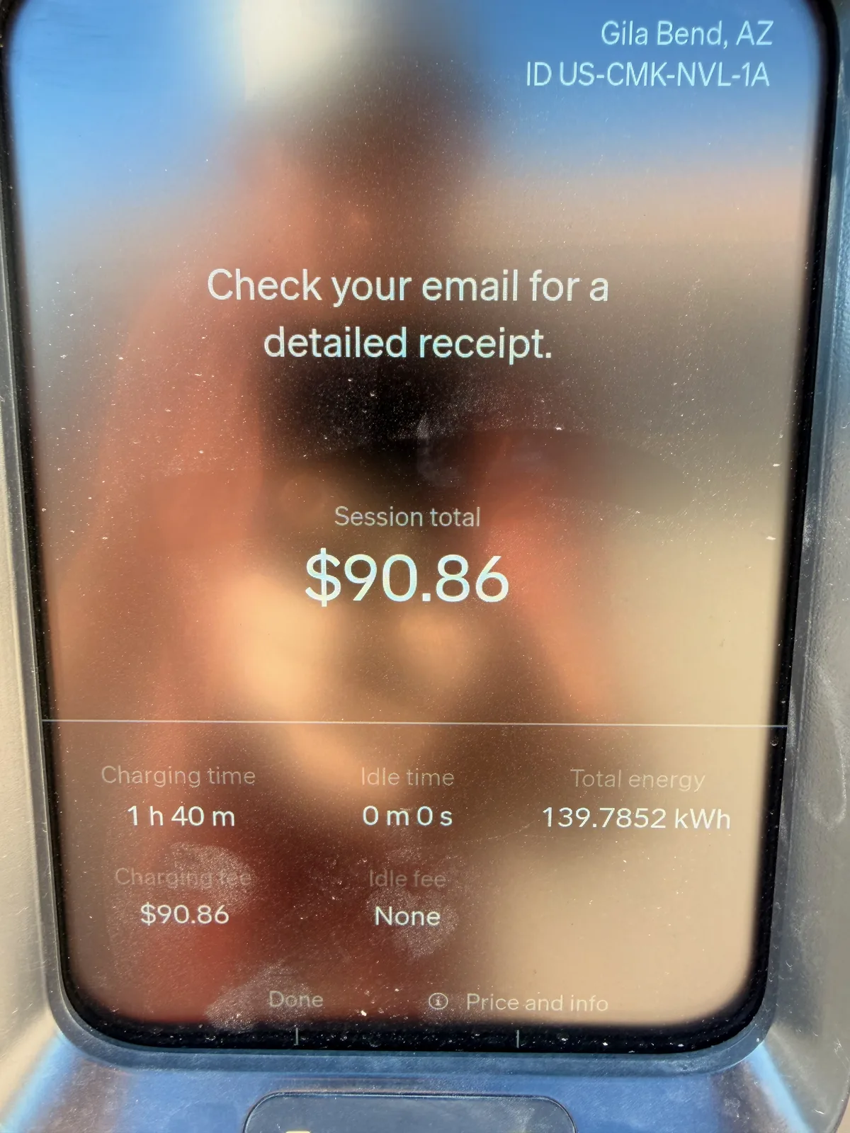 The Gila Bend RAN charger pedestal screen at 8:23 AM showing charger ID US-CMK-NVL-1A, a $90.86 session total, 1h 40m charging time, zero idle time, and 139.7852 kWh total energy delivered, with the photographer faintly reflected on the glass