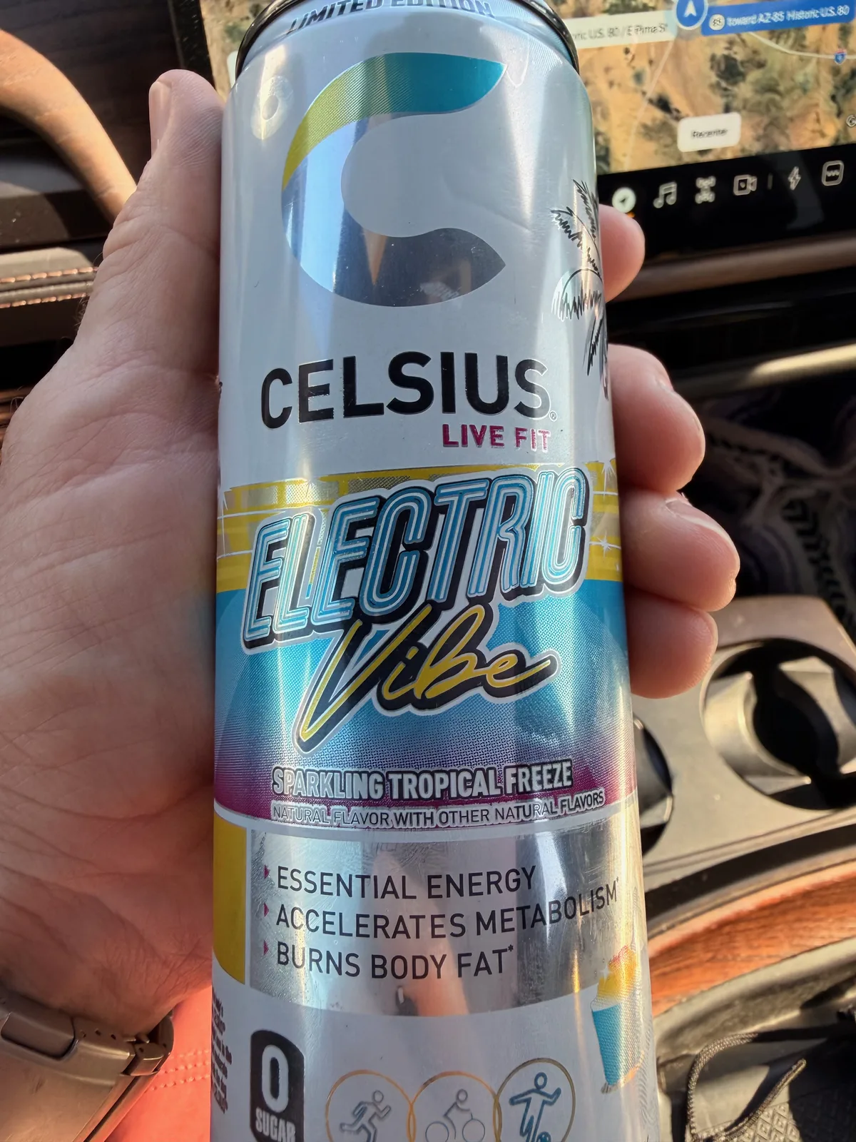 A Celsius Electric Vibe sparkling tropical-freeze energy drink can held up in the Rivian R1T cab with the AZ-85 highway and Pima Street signage visible on the center display behind it