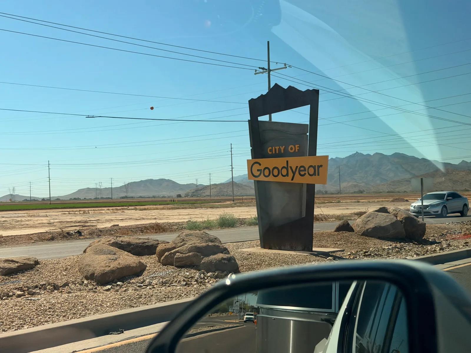 A "City of Goodyear" wedge-shaped sign on a roadside median against the Estrella Mountain range, viewed from the passenger side of the Rivian R1T with the side mirror in the corner