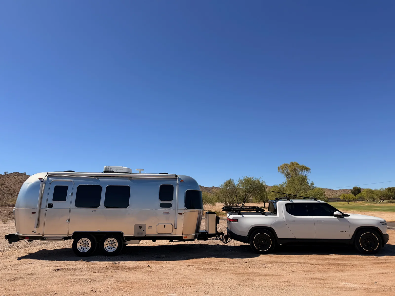 The silver Airstream International and white Rivian R1T parked broadside on a dirt pullout at Estrella Mountain Regional Park, with a low desert hill, sparse mesquite, and a strip of green park grass behind them under a deep blue Arizona sky