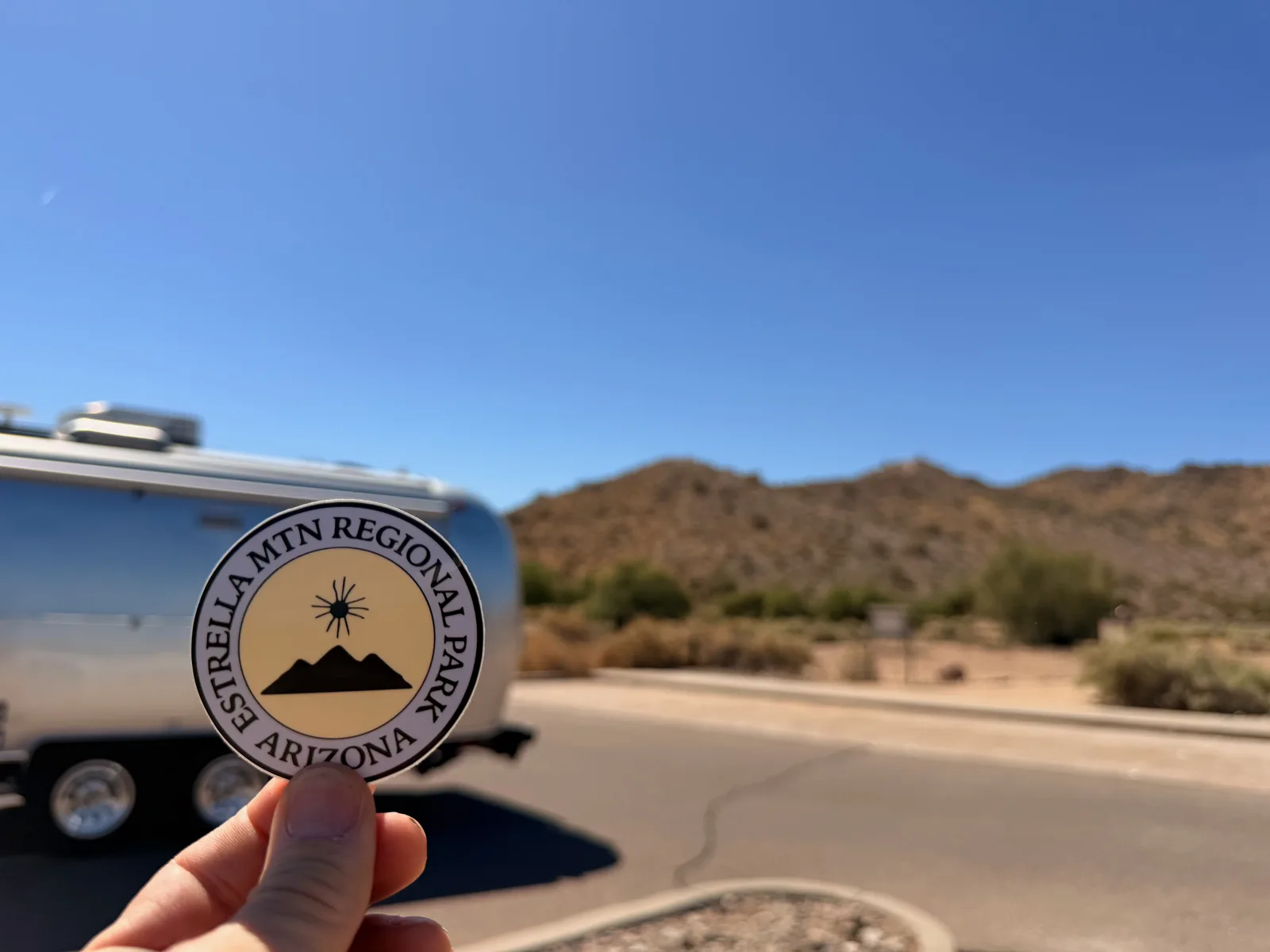 A round Estrella Mountain Regional Park Arizona souvenir sticker held up against the silver Airstream and the desert hills in soft focus, the park logo showing a sunburst behind two mountain peaks