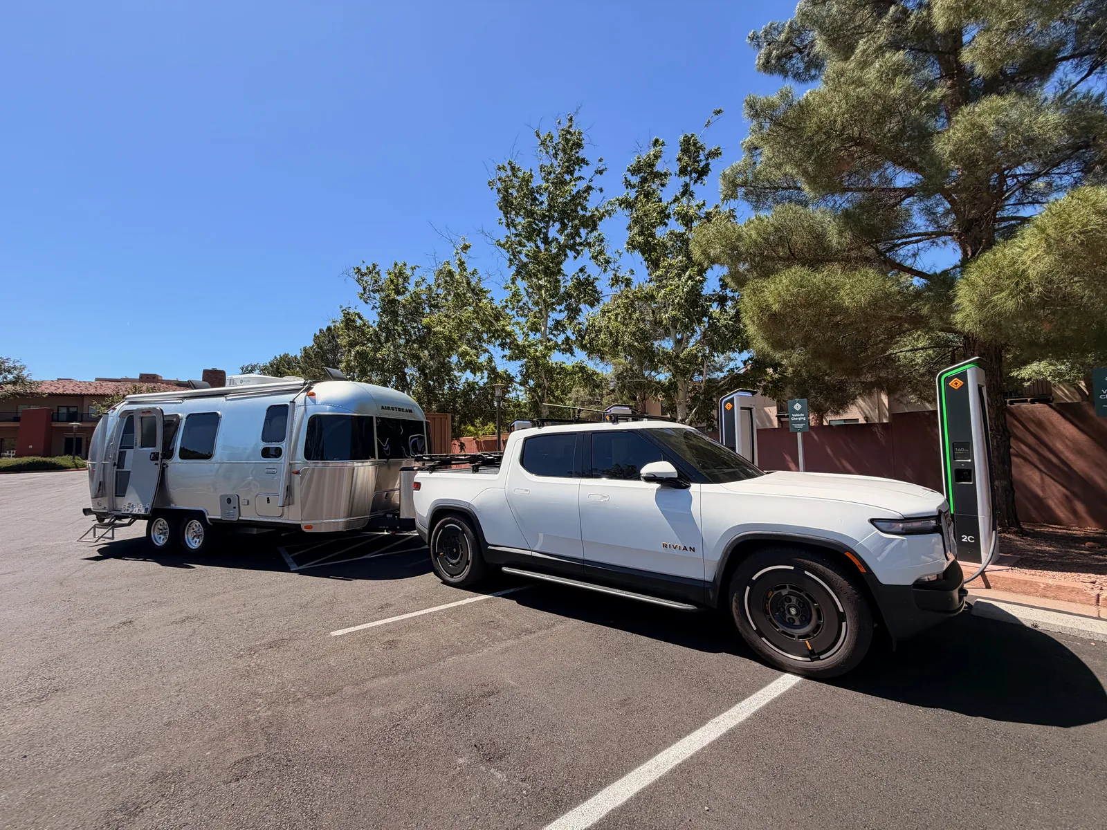 The white Rivian R1T and silver Airstream International parked at a green Rivian Adventure Network pedestal in the Village of Oak Creek, Sedona, framed by pine trees and adobe-tile rooflines under a deep blue Arizona sky