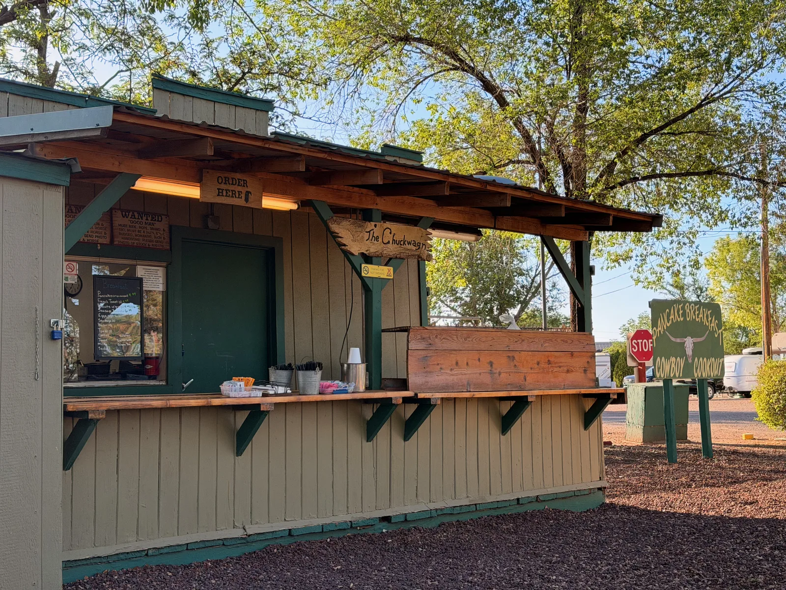 The Chuckwagon breakfast bar at the Holbrook KOA, a small wooden order-window stand with a Pancake Breakfast Cowboy Cookout sign next to it, cypress trees in the background under morning light