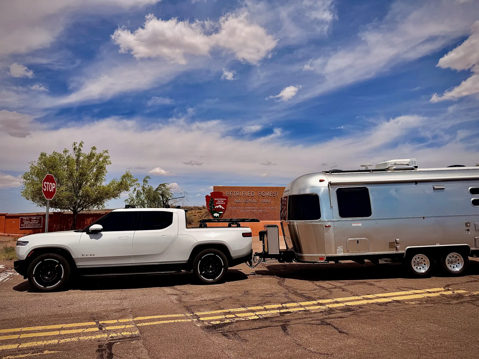 The Rivian R1T hitched to the silver Airstream International parked at the brown "Petrified Forest National Park" entrance sign with a stop sign and a small National Park Service kiosk behind, under a bright Arizona sky with scattered cumulus clouds