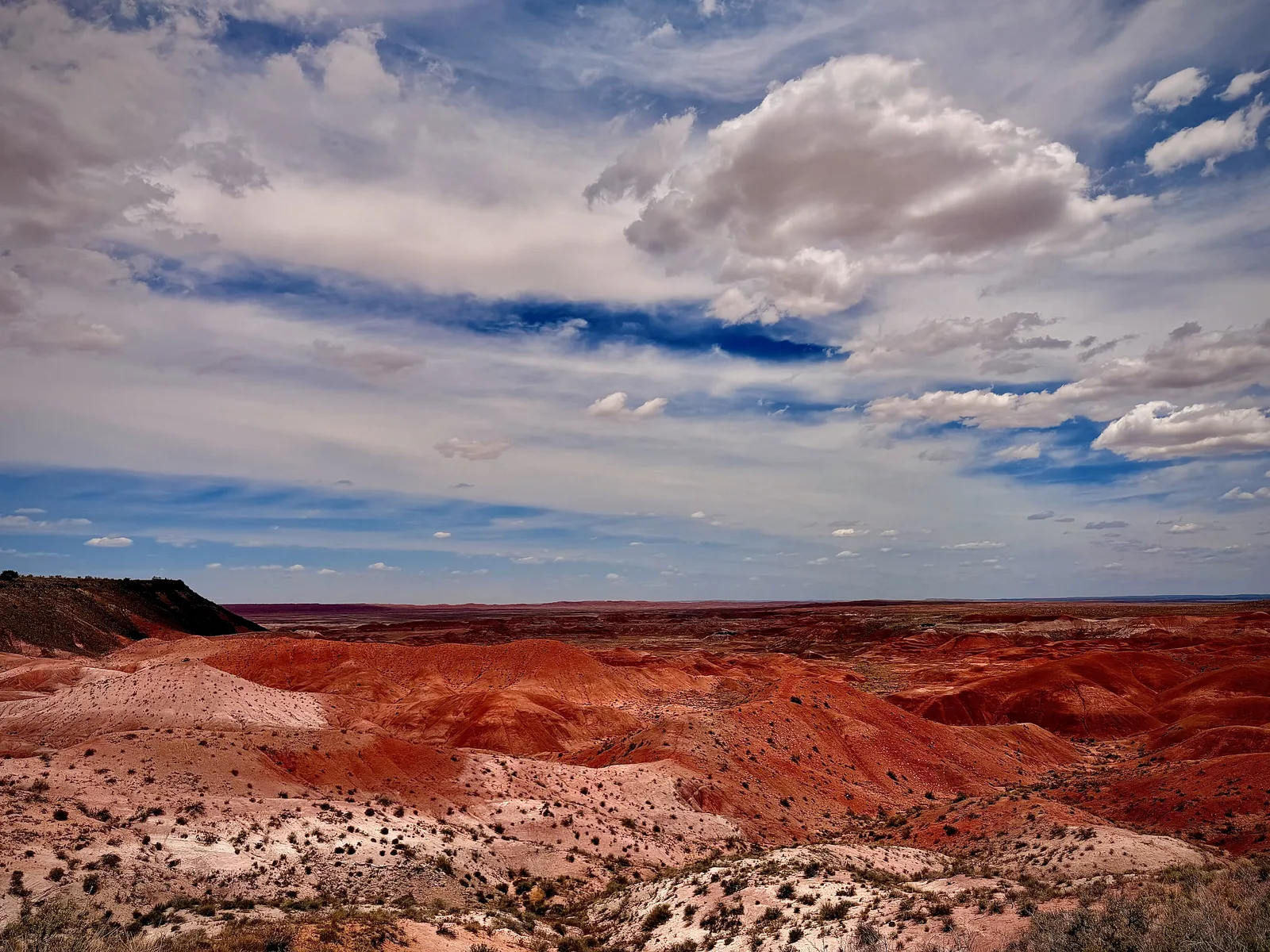 A wide Painted Desert vista from one of the rim overlooks, layered red and pink bentonite hills receding to the horizon under a deep blue sky with scattered white clouds, no people or vehicles in frame