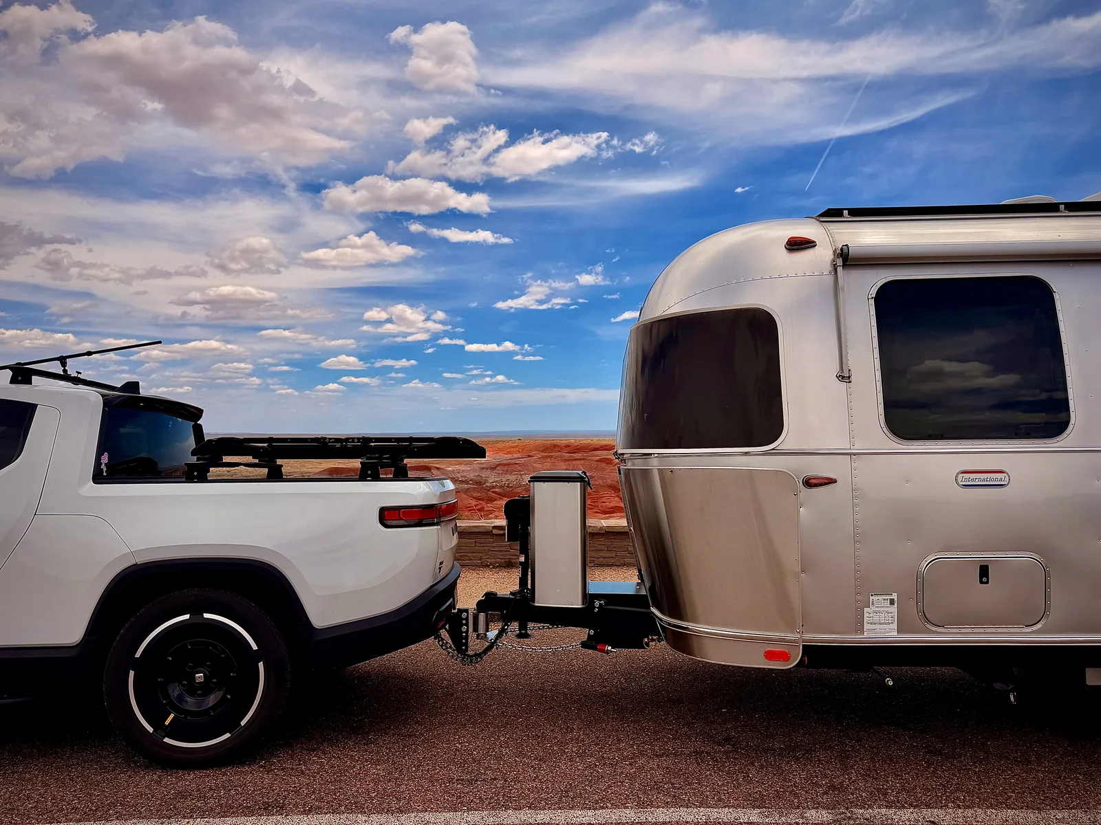Side view of the white Rivian R1T with roof rack hitched to the silver Airstream International, parked at a Painted Desert overlook with the rust-red badlands visible through the gap between the truck and the trailer