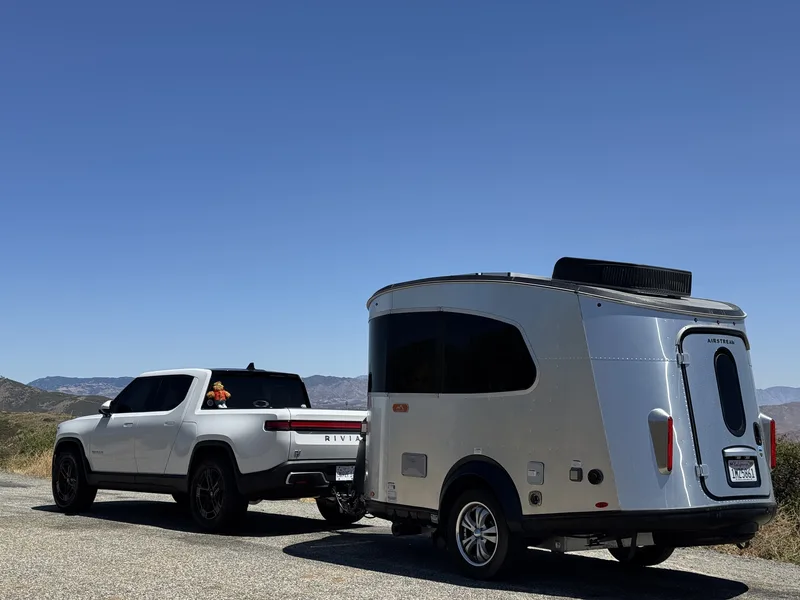 The Gen-1 R1T "Pablo Eskobear" hitched to the 16-foot Airstream Basecamp at a forested campsite outside Julian CA, the truck and trailer parked under tall pines on pine-needle ground