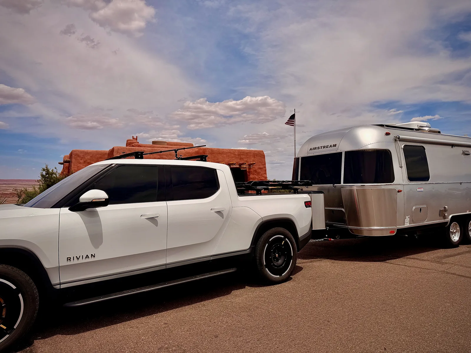 The white Rivian R1T and silver Airstream parked at the Painted Desert Inn, a 1937 adobe National Historic Landmark with a flag pole at the back, against the layered red Painted Desert and a sky full of cumulus