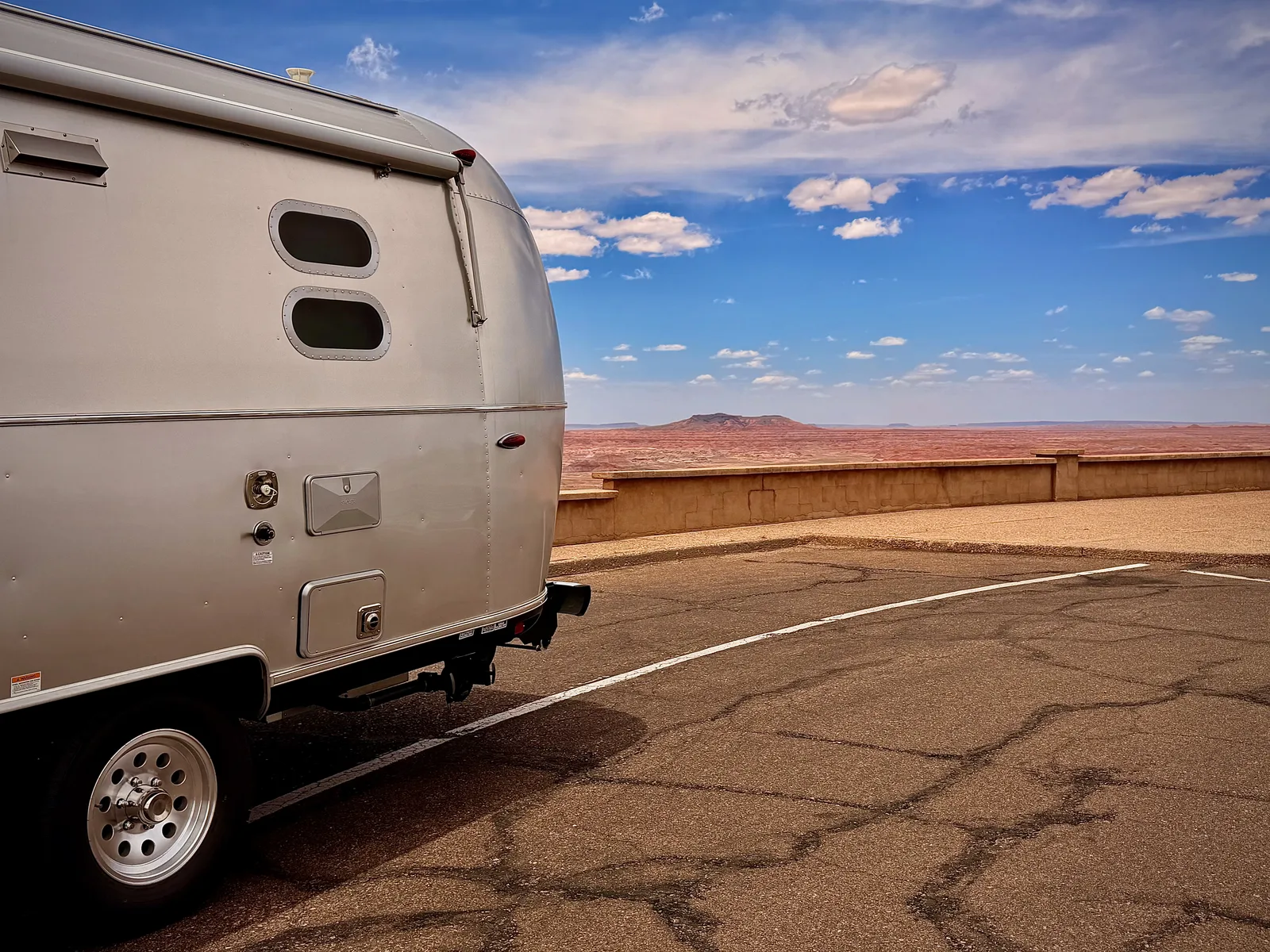 Rear angle of the silver Airstream parked at a Painted Desert overlook with the layered red sandstone hills and a small mesa in the distance under a partly-cloudy sky