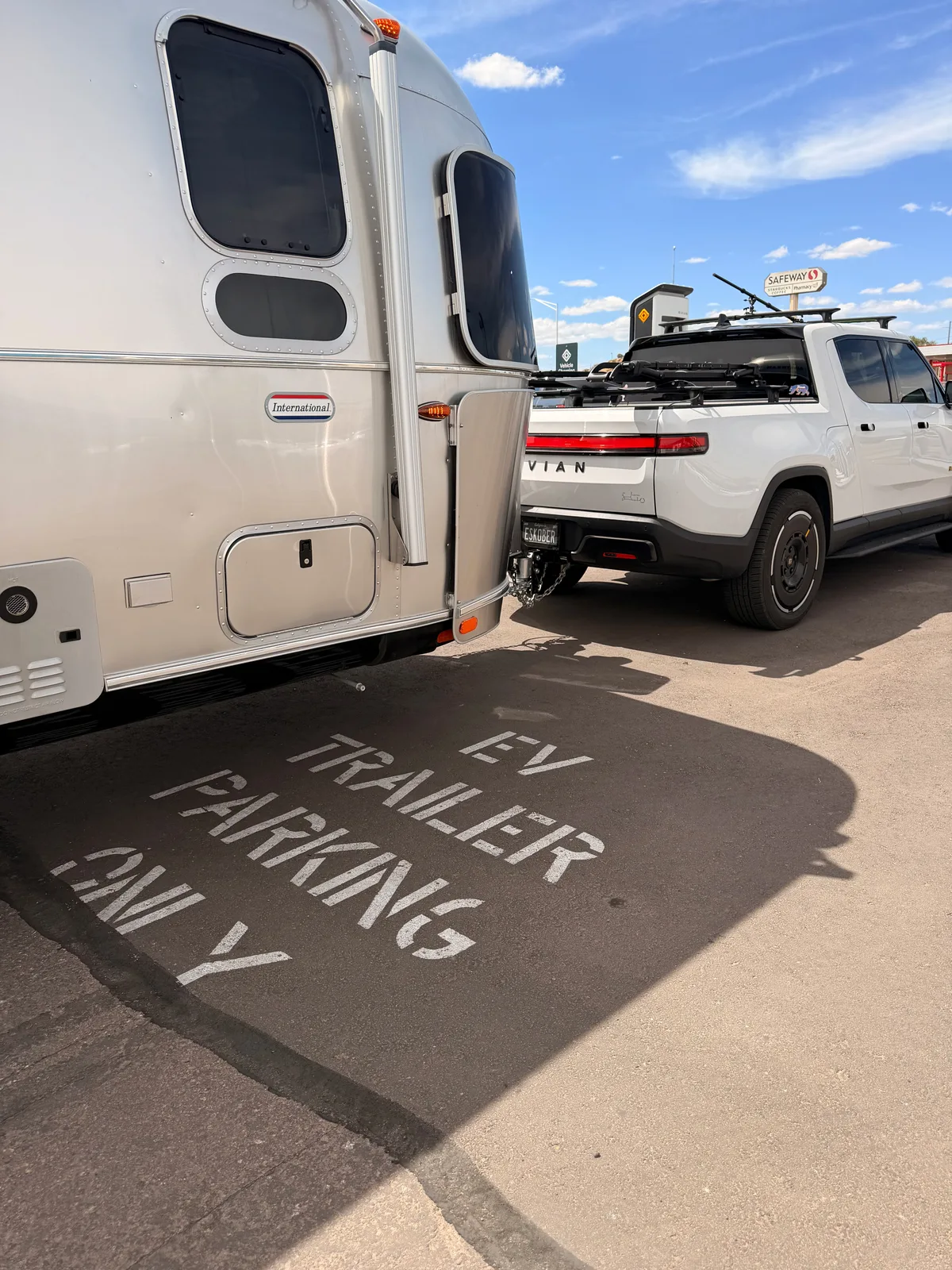 The white Rivian R1T hitched to the silver Airstream International parked over a yellow stenciled "EV TRAILER PARKING ONLY" marking on the asphalt at the Gallup Rivian Adventure Network site behind the Safeway, late-afternoon sun