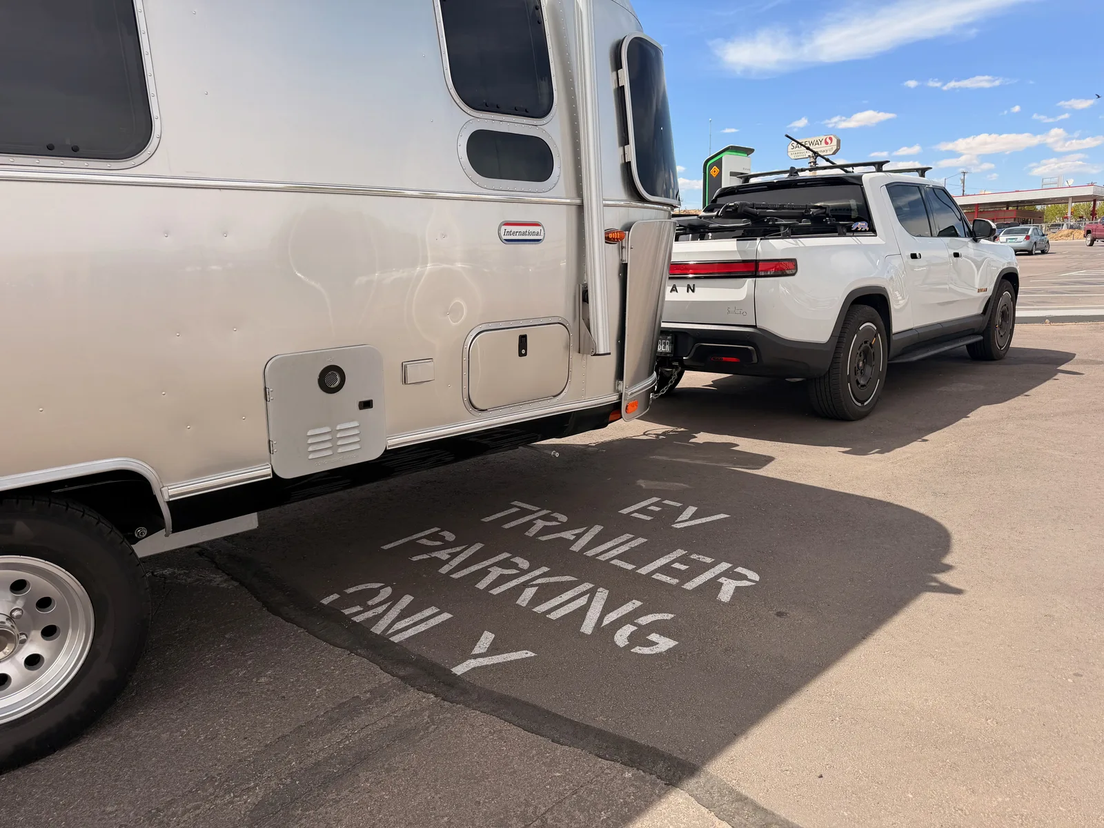 Wider landscape view of the white Rivian R1T and silver Airstream International parked at the Gallup RAN trailer-marked stall, with the tan RAN pedestal at left and the Gallup mesa skyline behind