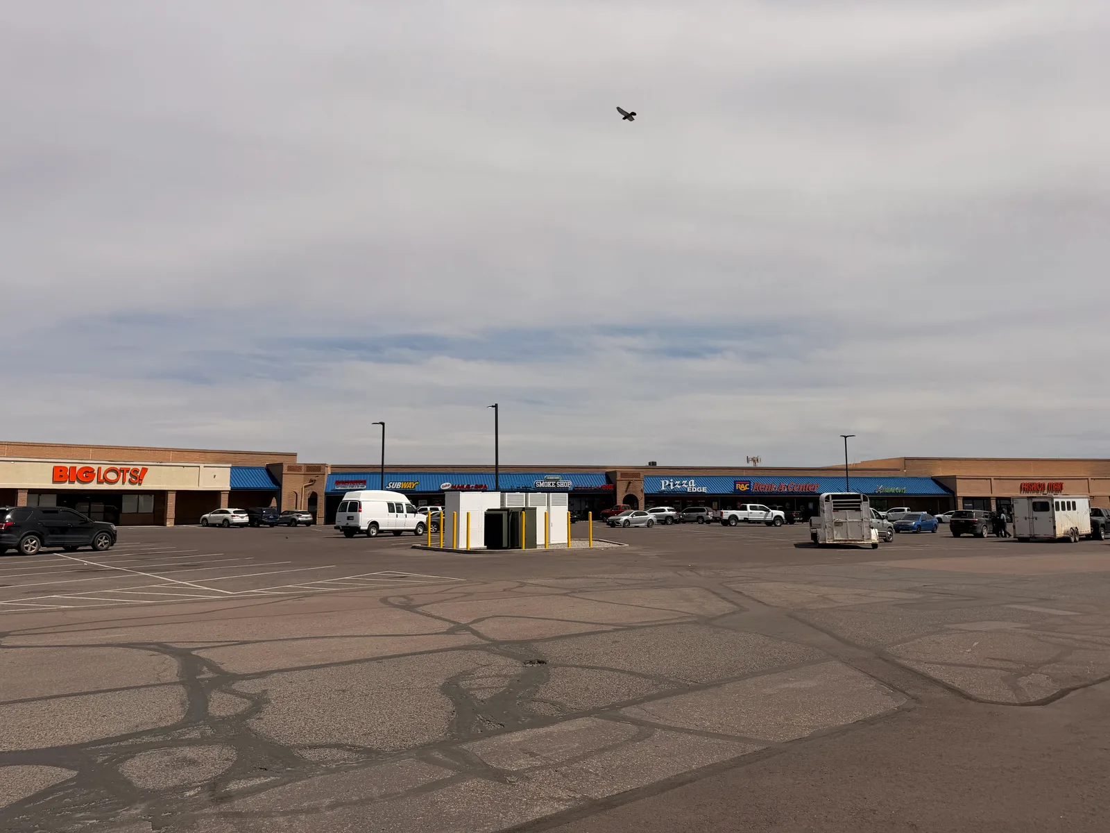 A wide view of the strip-mall parking lot behind the Gallup Safeway with the white Rivian R1T and silver Airstream parked at the Rivian Adventure Network site, surrounding storefronts including a Big Lots and Family Dollar in the background