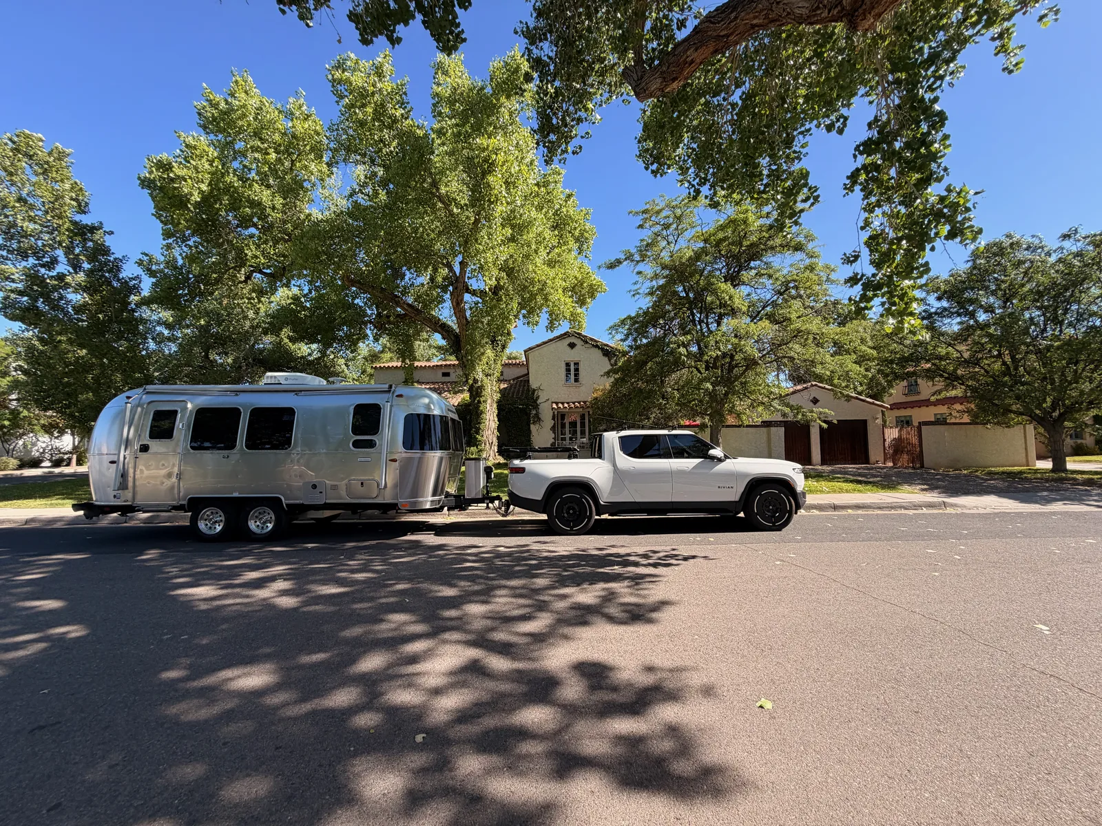 The Rivian R1T and silver Airstream International parked at the curb in front of the Jesse Pinkman house at 322 16th St SW, a Spanish Revival single-story with terra-cotta tile roof, ivy on the side wall, and a large cottonwood tree shading the driveway