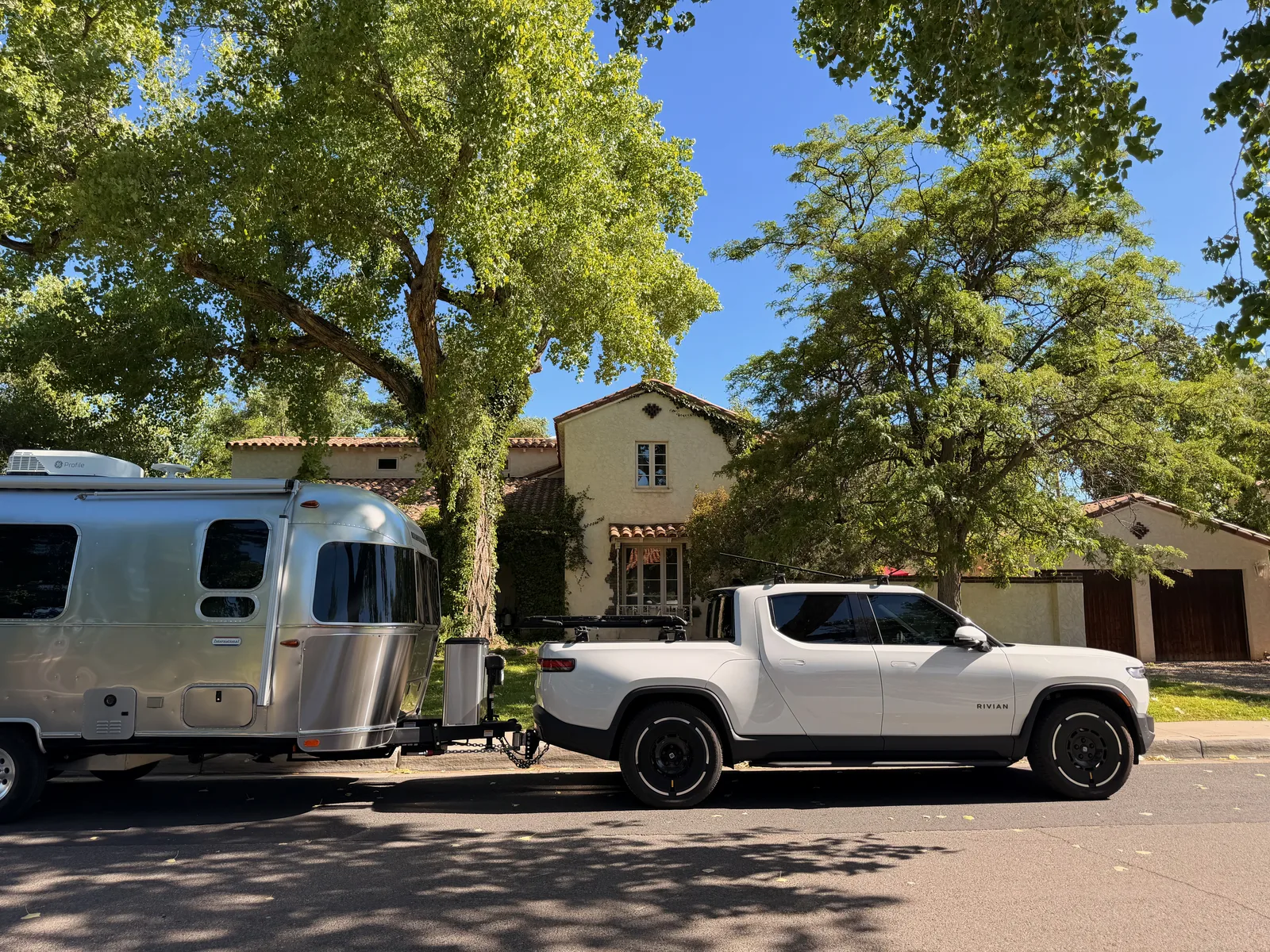 Closer angle of the rig parked at the curb at 322 16th St SW, the front quarter of the Rivian R1T visible alongside the silver Airstream, the Spanish Revival house and terra-cotta roof behind under bright morning light