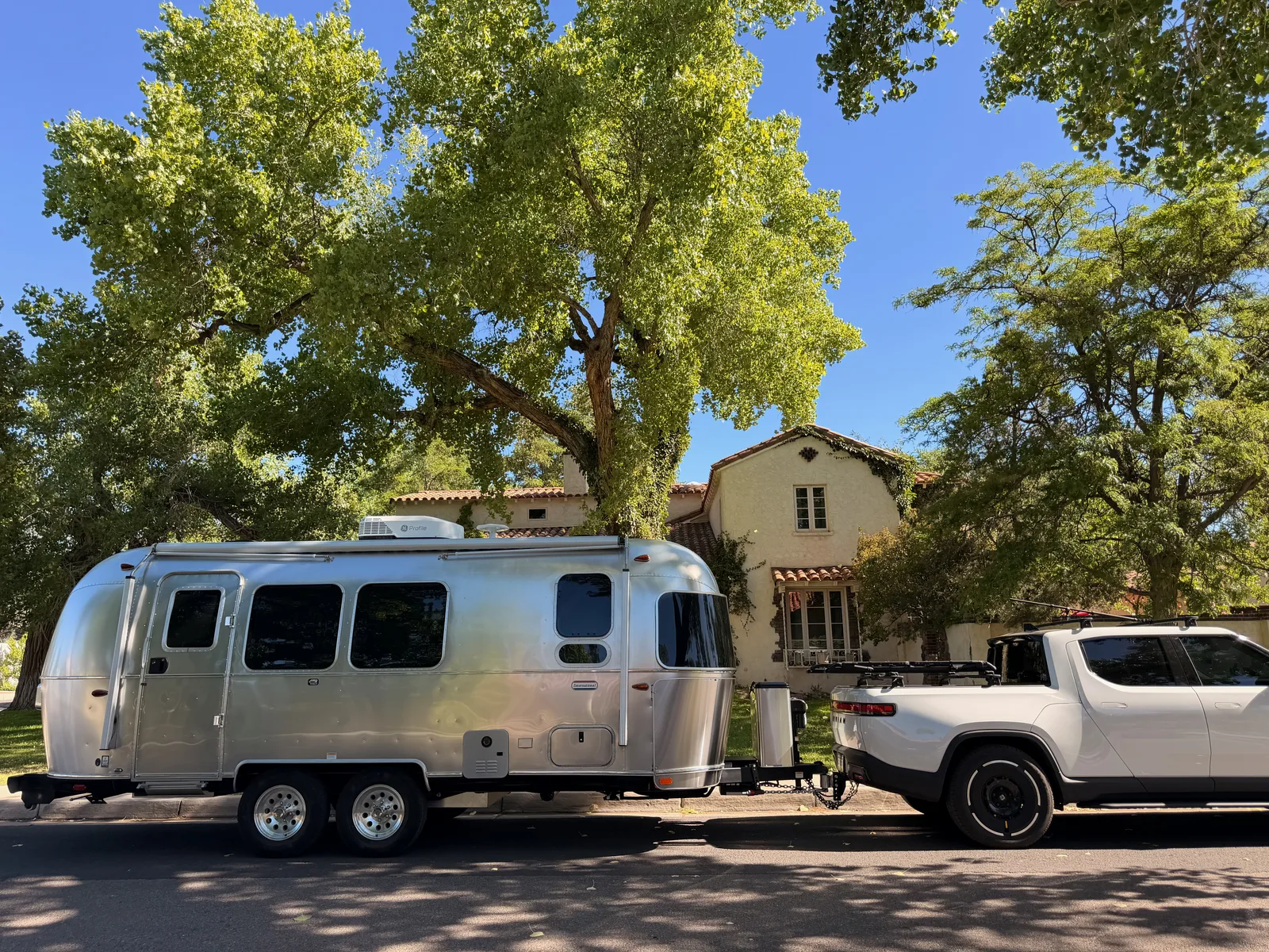 Wide street view of the Rivian R1T pulling the silver Airstream past 322 16th St SW, the Jesse Pinkman house framed by mature cottonwoods, terra-cotta tile roof and stucco walls in late-morning light