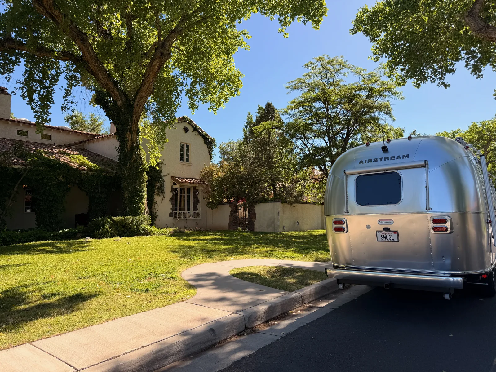 Three-quarter view of the rear of the silver Airstream International with California SMUGL plate parked in the street, the Jesse Pinkman house in the Spanish Revival style at left, ivy on the side wall, classic Breaking Bad framing