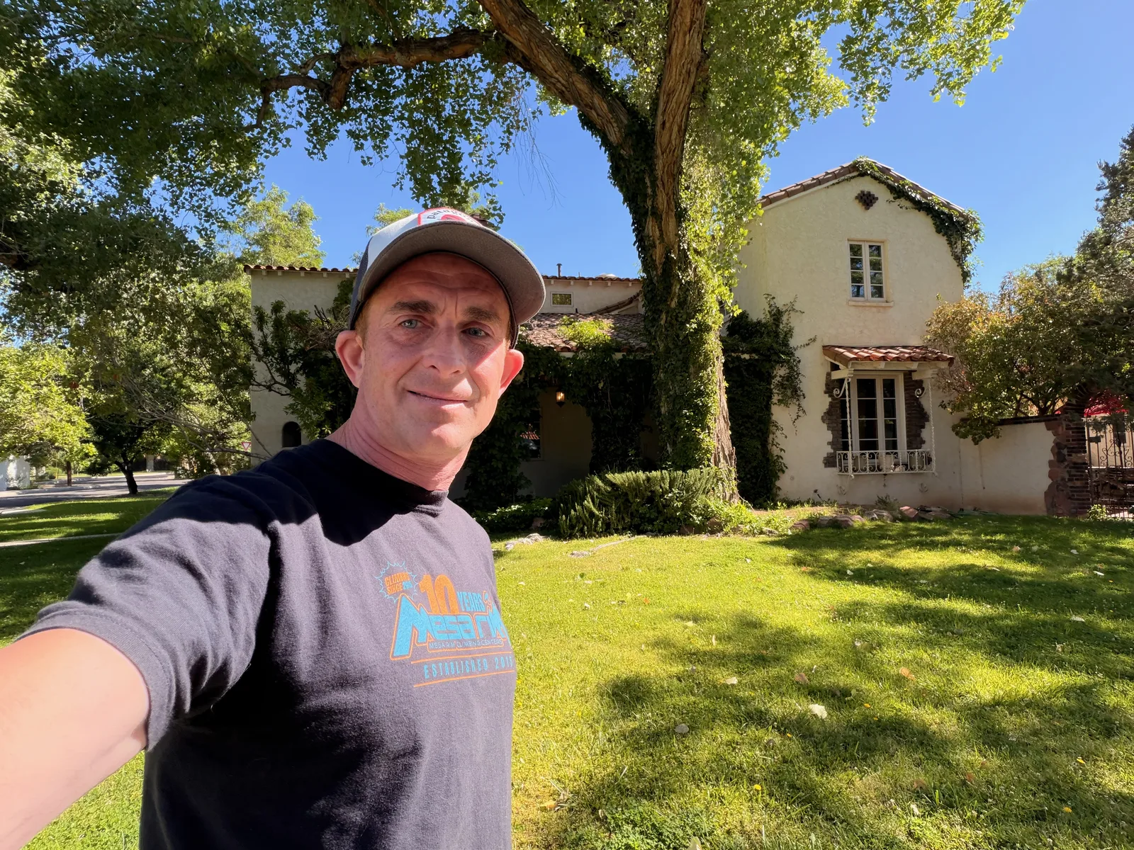 Eric (the author) standing in the front yard of the Jesse Pinkman house at 322 16th St SW, wearing a Mesa Rim 10 Years t-shirt and a baseball cap, the Spanish Revival house with terra-cotta tile roof and ivy framed behind him under a blue New Mexico sky