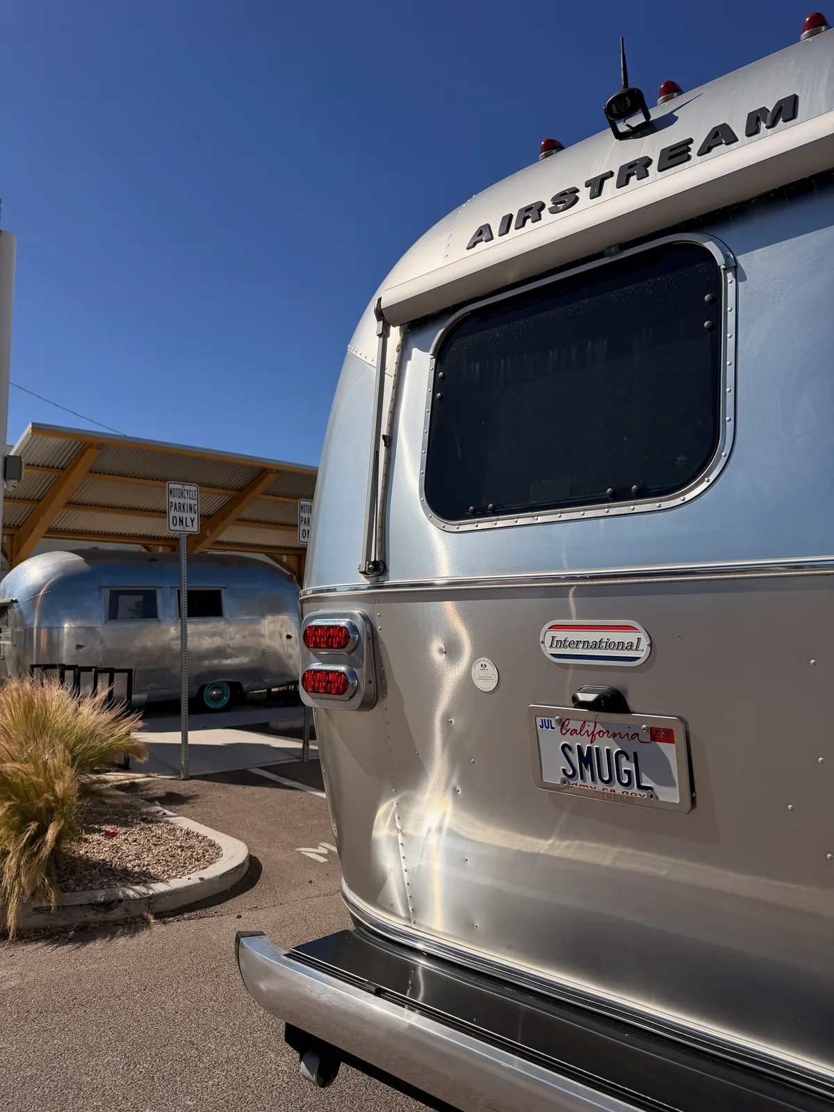 The rear of the silver Airstream International with California SMUGL plate parked next to a vintage 1960s polished-aluminum Airstream travel trailer displayed as public art under a canary-orange canopy, the contrast between new and old visible on the same lot