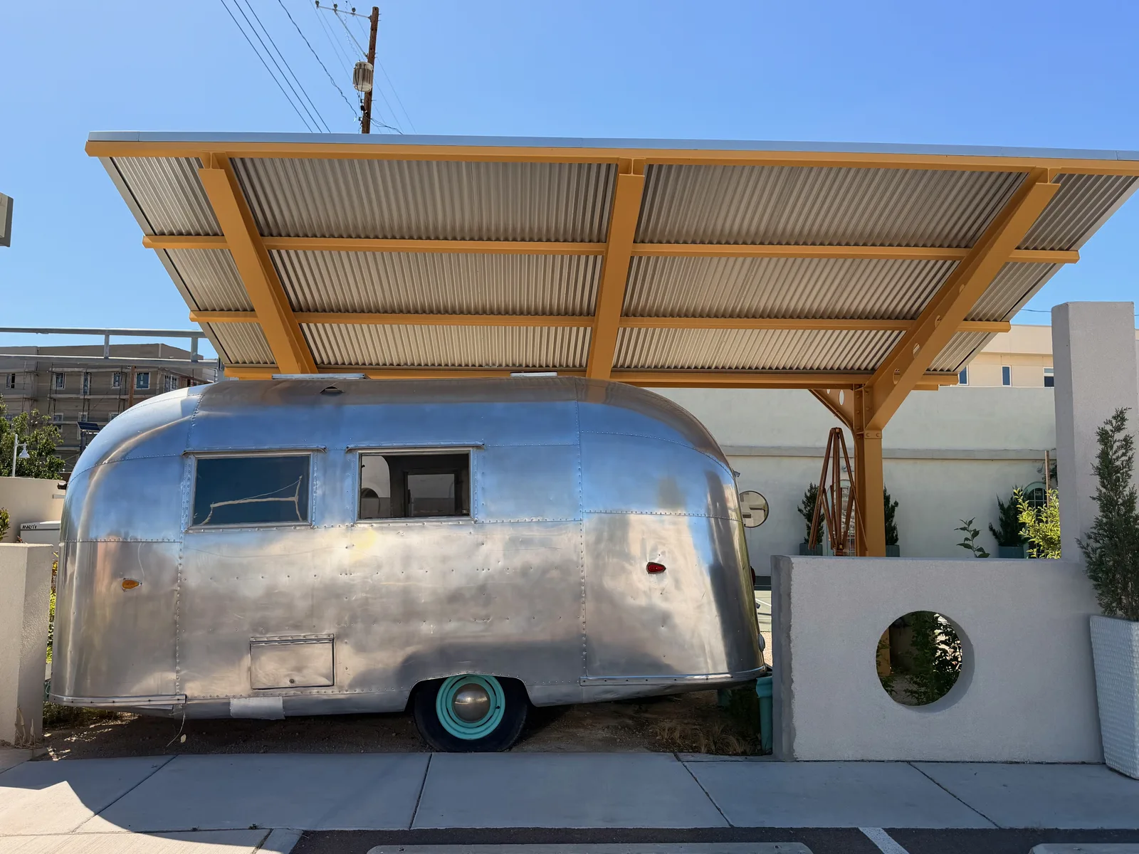 Wider view of the vintage 1960s polished-aluminum Airstream travel trailer under the canary-orange canopy with a porthole window in the white wall behind, a Route 66 era public art installation in Albuquerque
