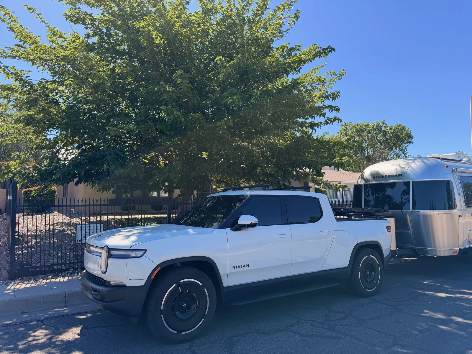 Front-three-quarter view of the Rivian R1T and Airstream parked along the curb at the Walter White house, the black wrought-iron fence and gravel landscaping of the property visible at left, midday sun and a deep blue New Mexico sky