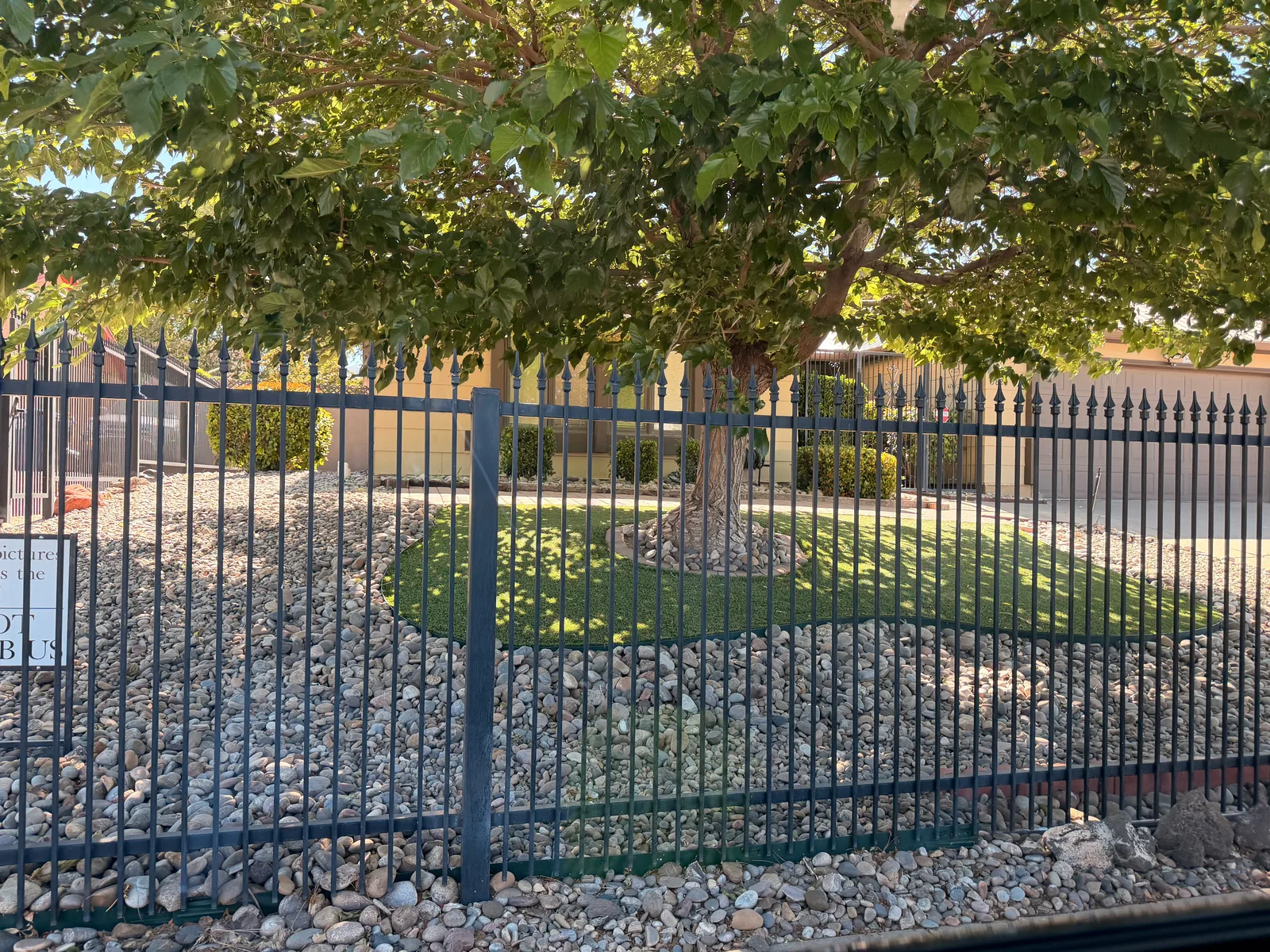 A "No Photographs" sign on the wrought-iron security fence at the Walter White house, with mature trees, gravel landscaping, and a glimpse of the iconic two-story brick home behind, the sign a result of years of Breaking Bad tourists overwhelming the property
