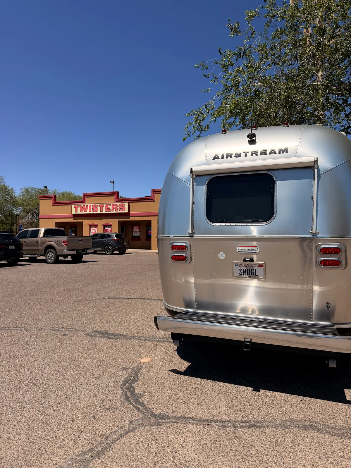 The rear of the silver Airstream International with California SMUGL plate parked in the Twisters Burgers and Burritos lot at 4257 Isleta Blvd SW, the rust-orange building with red TWISTERS signage visible across the lot under a deep blue New Mexico sky