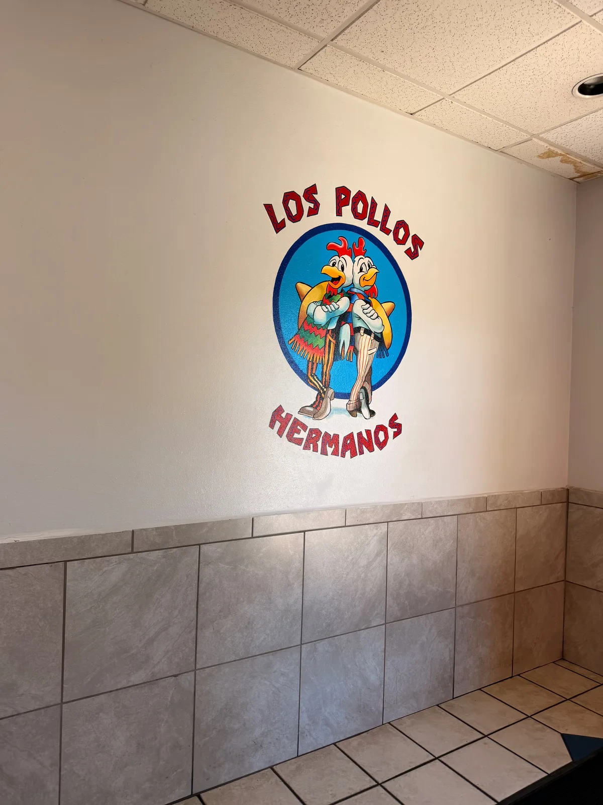 The interior chicken-couple LOS POLLOS HERMANOS mural inside the Twisters restaurant, two cartoon chickens in dresses and hats holding the logo on a white wall above a tan tile wainscot