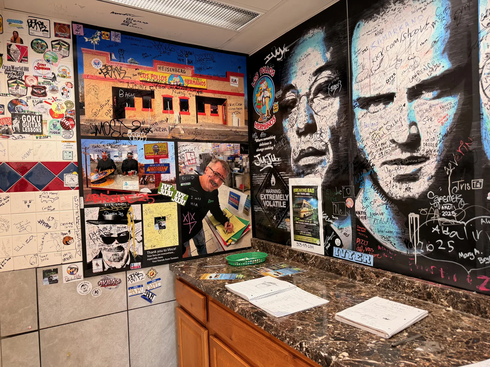 A back-counter shrine at Twisters covered in Breaking Bad memorabilia, a large painted portrait of Bryan Cranston as Walter White and Aaron Paul as Jesse Pinkman heavily signed in marker by fans, sticker-covered tile, a "Breaking Bad Extremely Volatile" sign, and a guestbook on the counter
