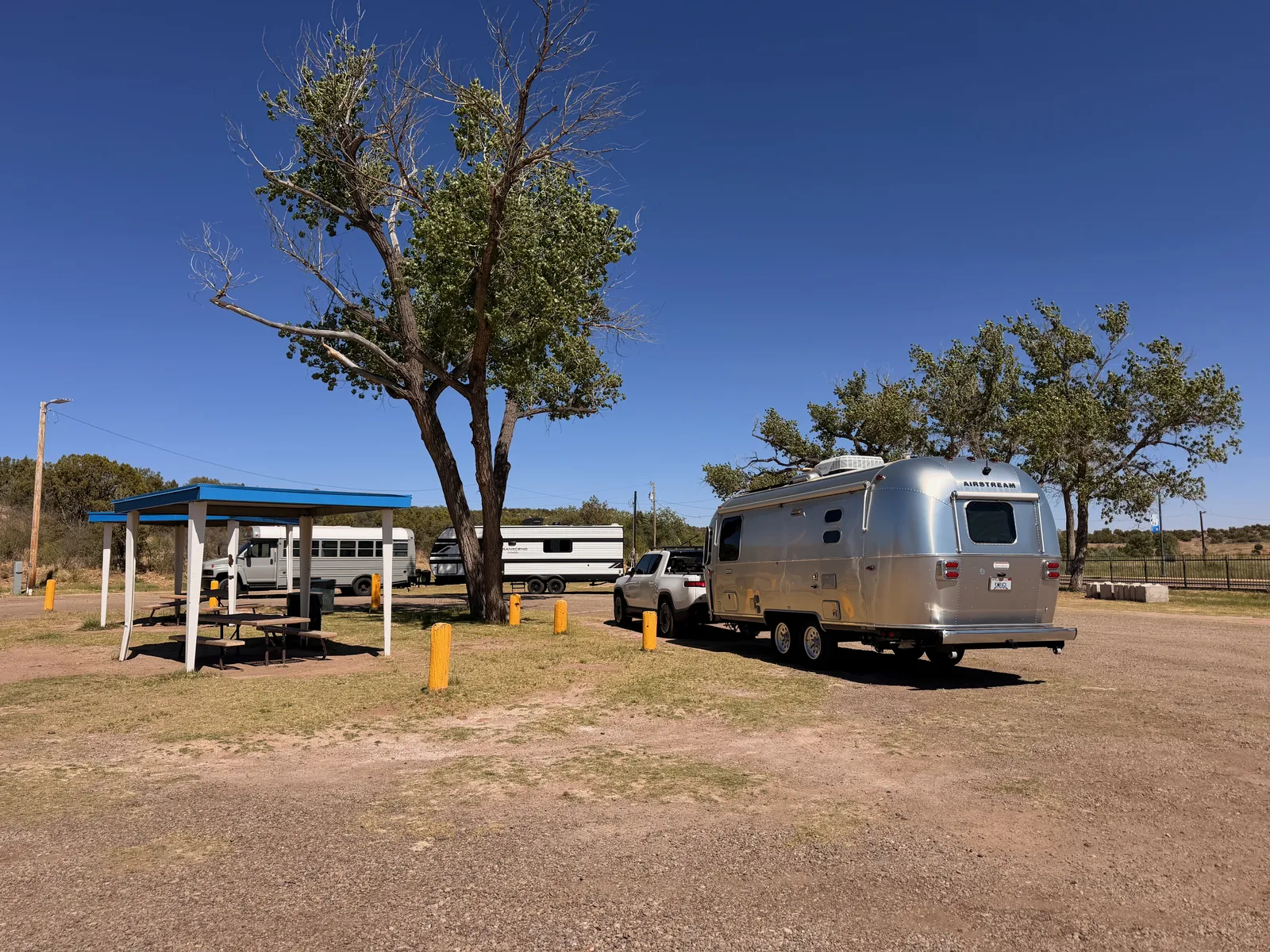 The white Rivian R1T hitched to the silver Airstream International parked in the Blue Hole lot at Santa Rosa NM, the park's blue picnic shelter at left, a cottonwood tree shading the gravel, and a horse trailer parked across the lot