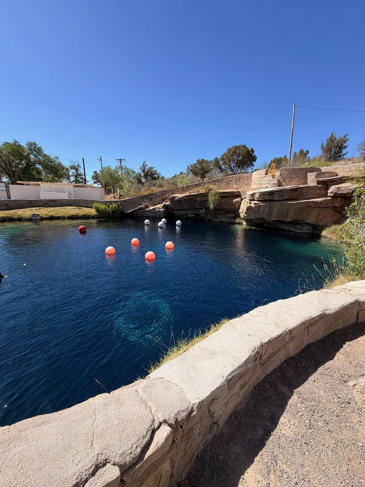 The Blue Hole at Santa Rosa NM, a crystal-clear deep-blue circular artesian spring with orange and pink safety buoys floating on the surface, sandstone rim walls and high-desert vegetation around the edge