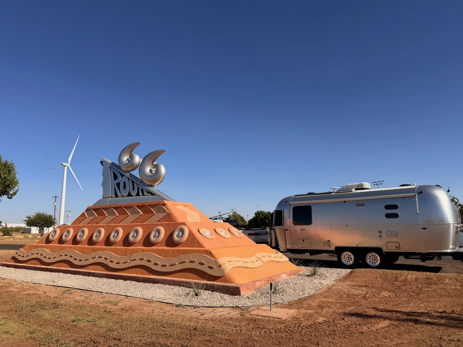 The Tucumcari Route 66 Monument, a giant orange-adobe pyramid sculpture with a steel ROUTE 66 cutout and chrome eye-like circles on top, the silver Airstream International parked alongside, a wind turbine spinning in the strong SW wind in the background