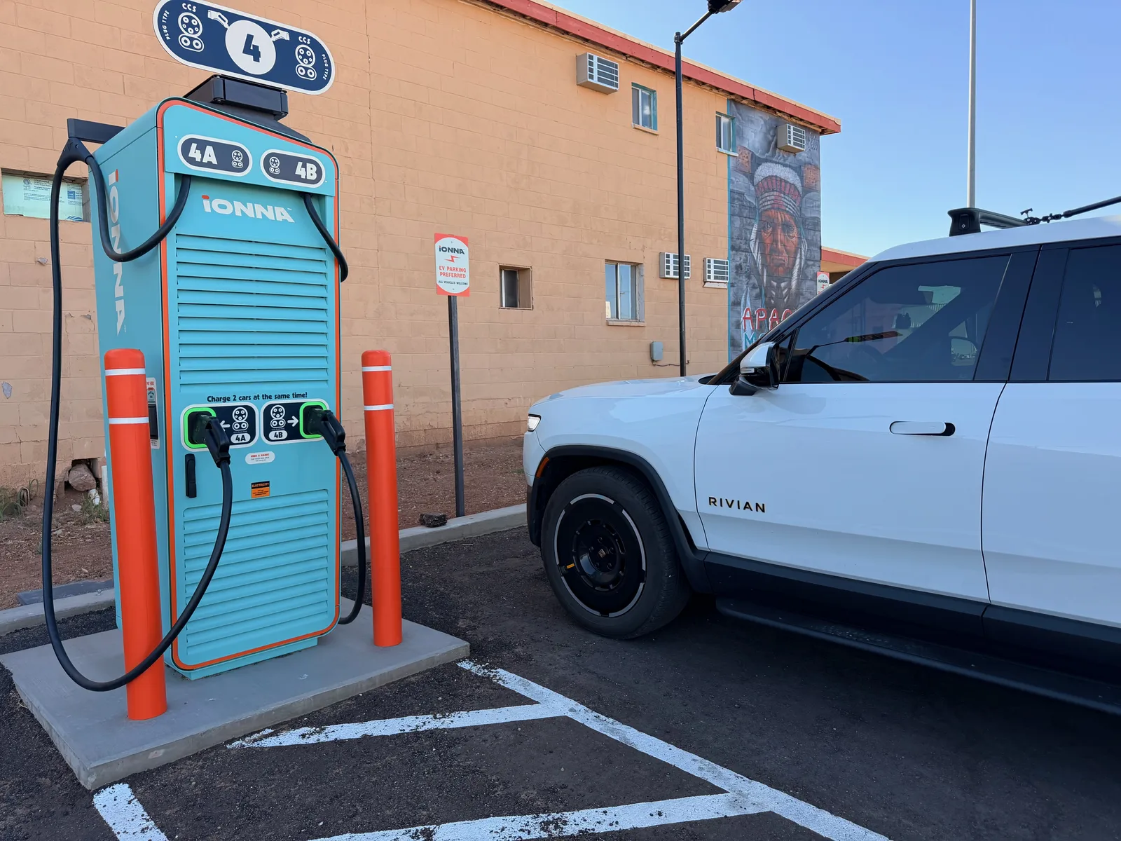 The white Rivian R1T parked at the teal-and-orange IONNA Rechargery pedestal in Tucumcari NM, dual cables for stalls 4A and 4B, a Native-American-chief mural on the side of the adjacent Apache Motel building visible behind, "Charge 2 cars at the same time" graphic on the cabinet