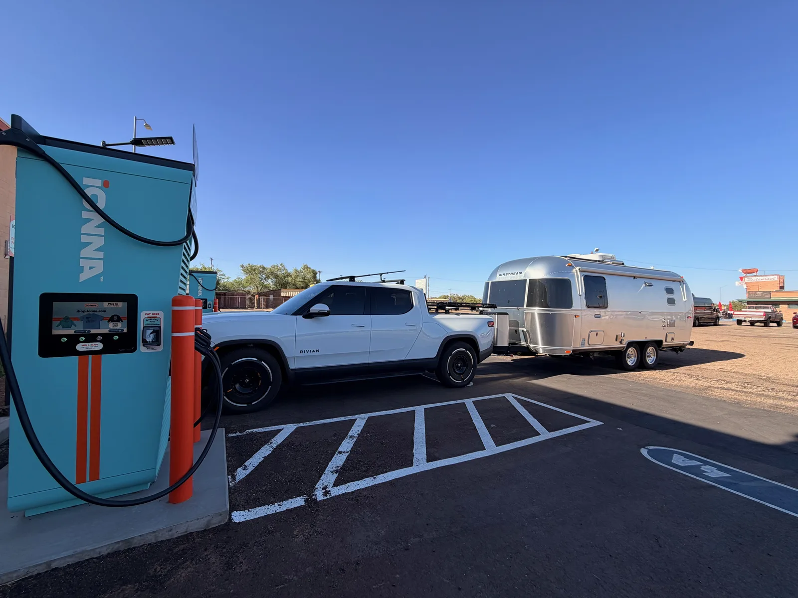 Three-quarter rear view of the white Rivian R1T plugged into the IONNA pedestal towing the silver Airstream, the IONNA wordmark on the cabinet at left, orange bollards protecting the pedestal, Route 66 motel signs visible in the distance down the boulevard at right