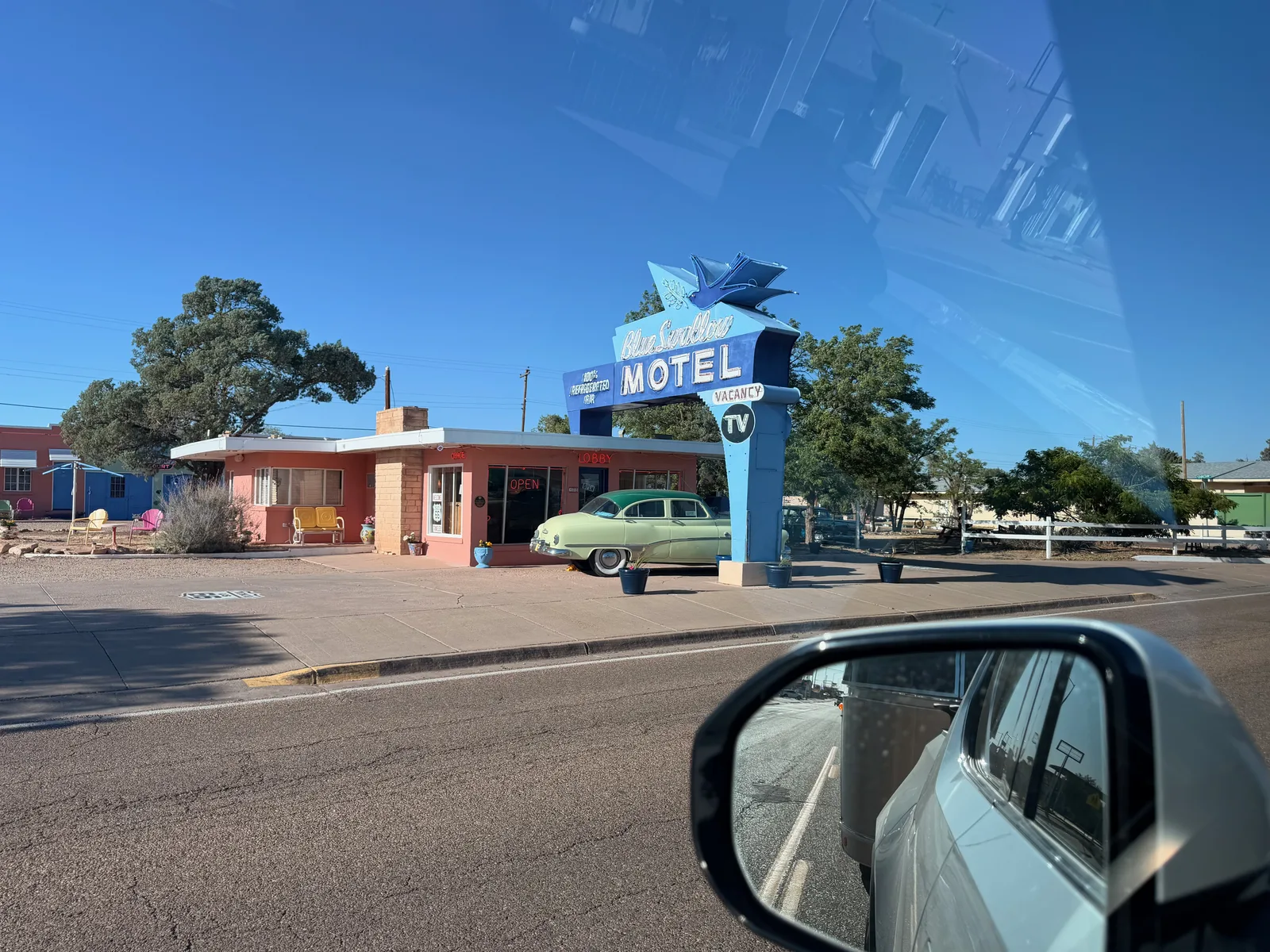 The Blue Swallow Motel in Tucumcari NM, the most intact 1939 Route 66 neon motel, shot from the side window of the moving Rivian, pink-coral stucco office with neon "Blue Swallow MOTEL" sign topped by a stylized blue swallow bird in flight, "100% AIRCONDITIONED" / "VACANCY" / "TV" panels, a period-correct 1951 Chevy sedan parked at the curb out front, the Rivian's side mirror visible at the bottom right