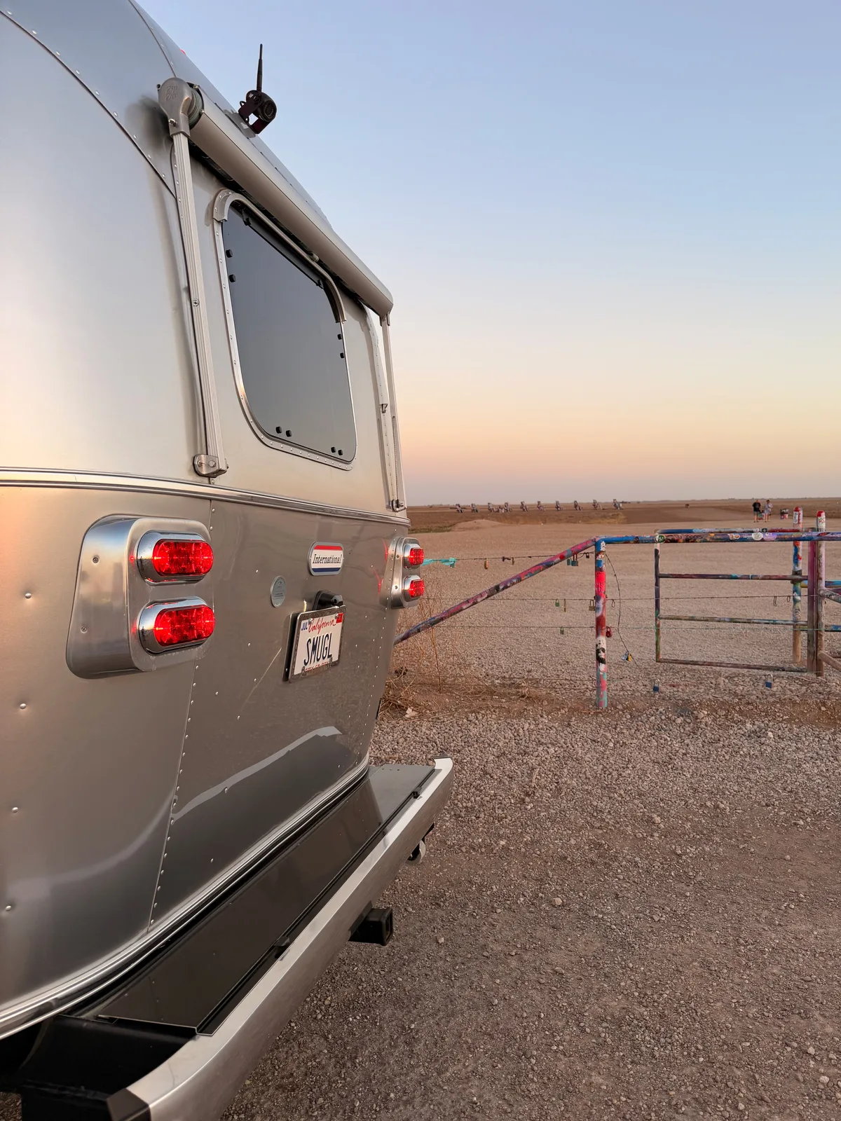 The rear of the silver Airstream International with the California SMUGL plate at Cadillac Ranch, the buried Cadillac sculptures lined up across the gravel field with their layered spray-paint graffiti catching warm sunset light, a graffiti-covered gate post in the foreground
