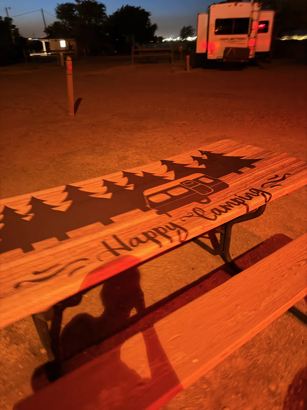 Night arrival at the Amarillo KOA Holiday, a wooden picnic table with a "Happy Camping" silhouette engraving in the foreground under warm campsite lighting, a neighbor's fifth-wheel partially visible, KOA power pedestal lit at left