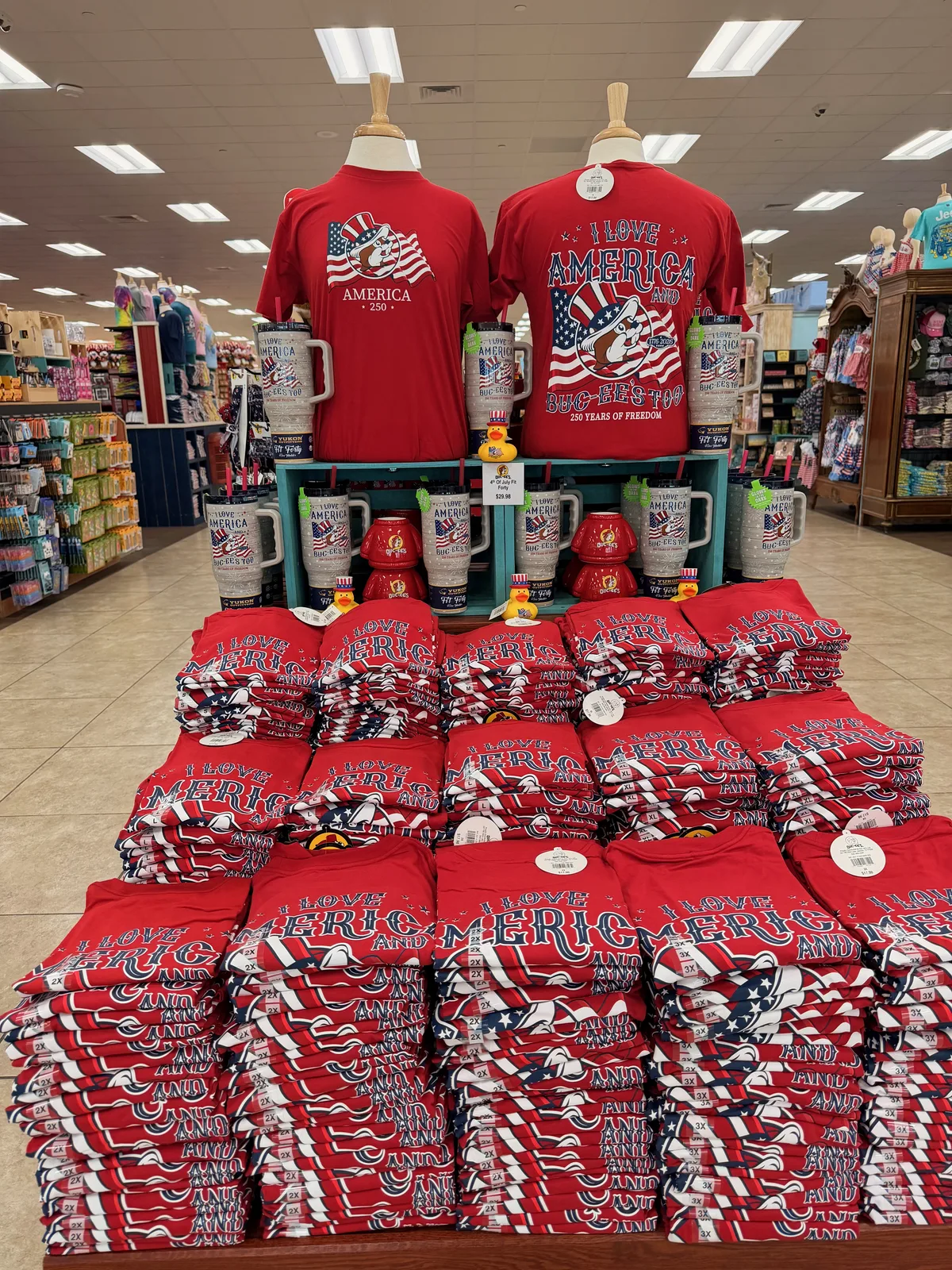 Inside the Buc-ee's Amarillo store, a tiered display of red "I LOVE AMERICA, BUC-EE'S" t-shirts arranged on a teal-blue circular table with branded mugs and rubber-duck figurines, the rest of the apparel section visible behind