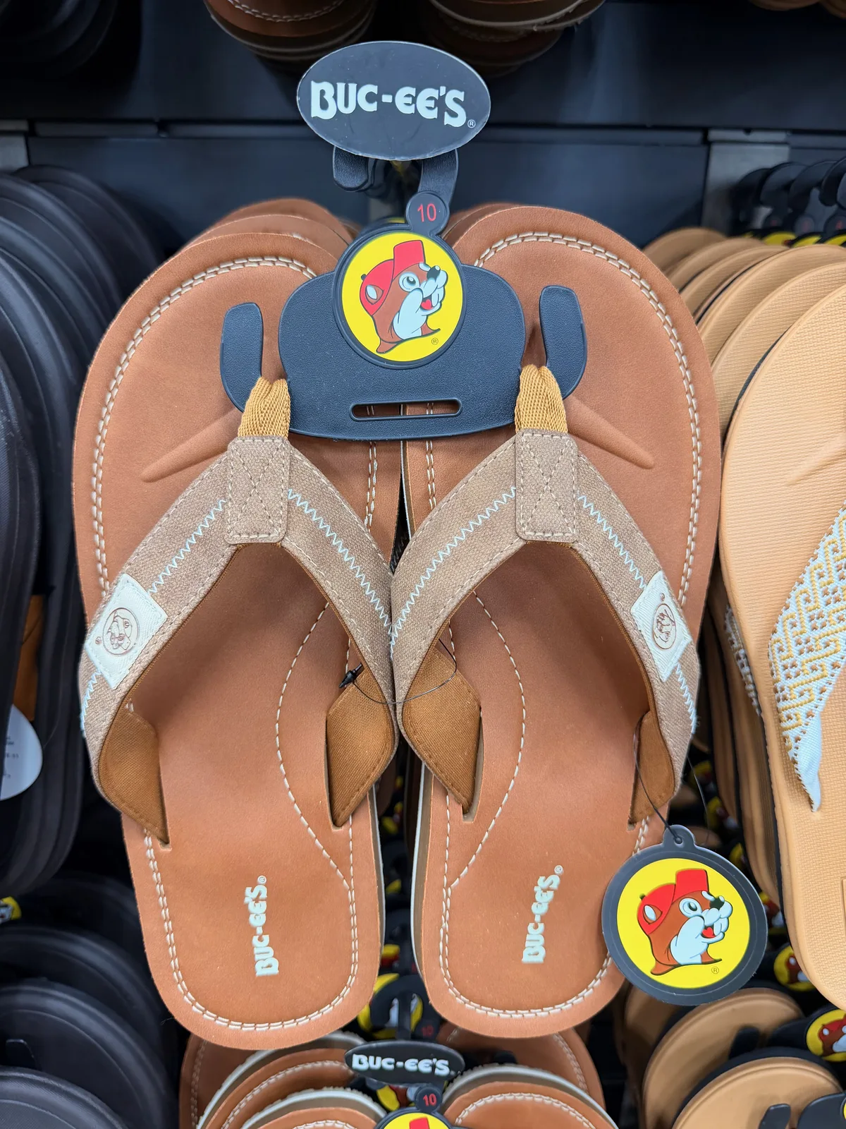 A pair of Buc-ee's-branded leather flip-flops in tan with the smiling Buc-ee beaver mascot logo on the strap, photographed on the in-store hanger display with the Buc-ee's wordmark tag above