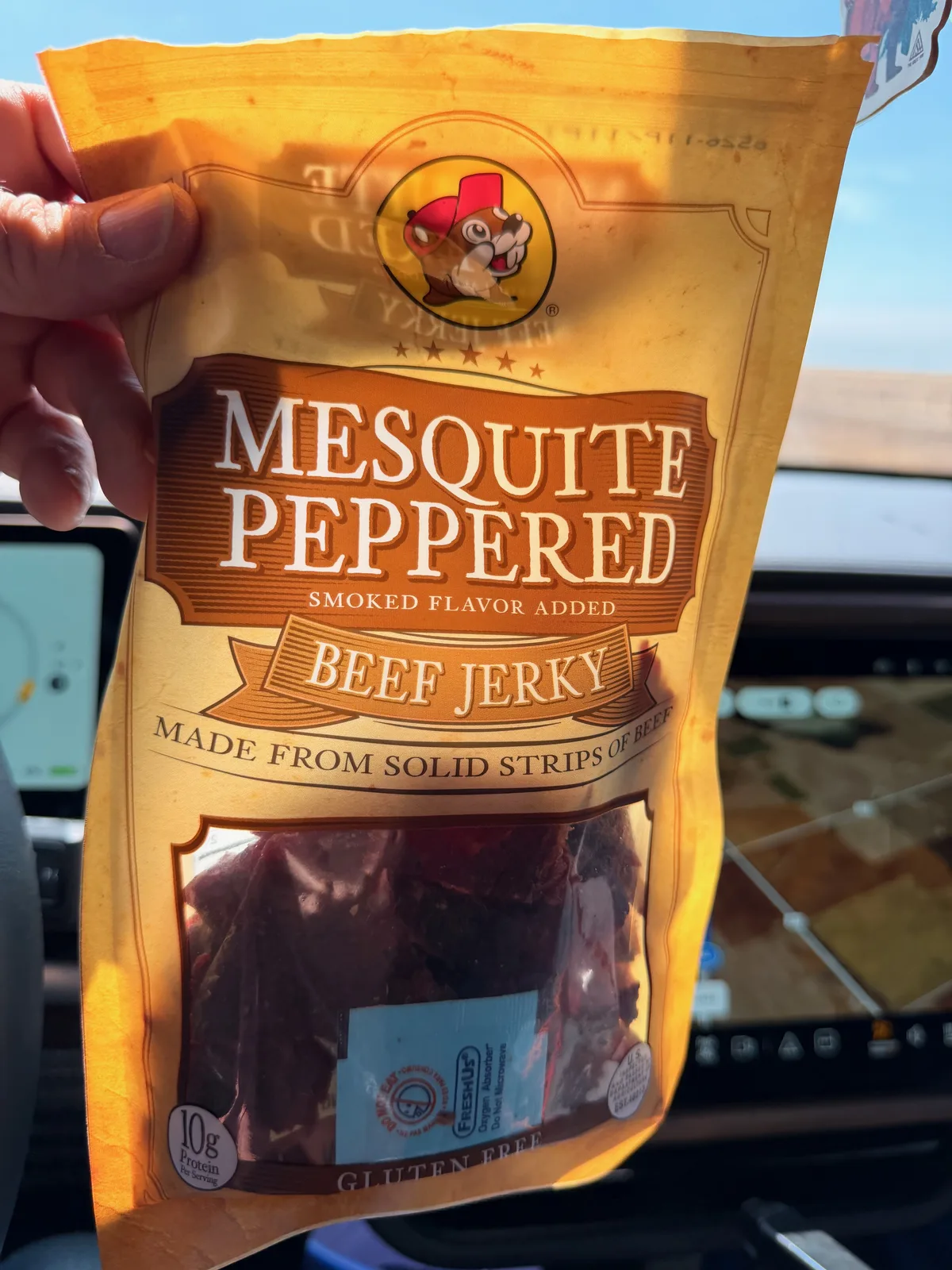 A Buc-ee's-branded "Mesquite Peppered Beef Jerky" bag held up in the cabin of the Rivian R1T while driving east on I-40, the action camera on the dash visible and the Texas Panhandle landscape blurring past through the windshield