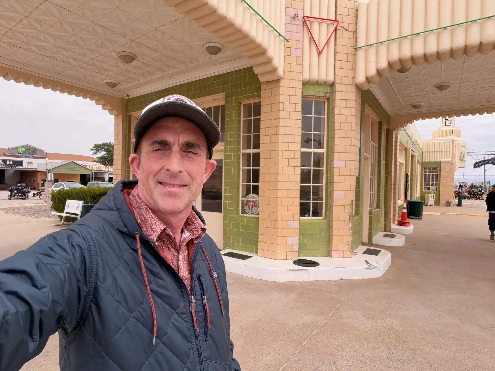 Eric standing under the green-glazed Art Deco tile canopy of the U-Drop Inn in Shamrock TX, the cream and green Art Deco facade behind him, a Holiday Inn Express visible across the street, overcast Texas sky