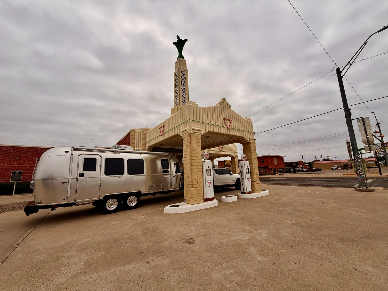Wide-angle exterior of the U-Drop Inn from the parking lot, the silver Airstream and white Rivian R1T tucked under the Art Deco canopy with the cream Conoco tower above and the vintage gas pumps in the foreground, downtown Shamrock TX visible across the street