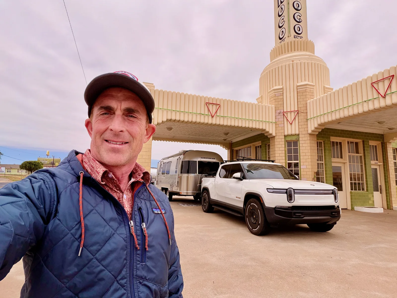 Eric selfie in front of the U-Drop Inn in a blue puffy jacket and Mesa Rim cap, the cream Art Deco Conoco tower with vertical signage rising behind, the rig parked under the canopy in the background