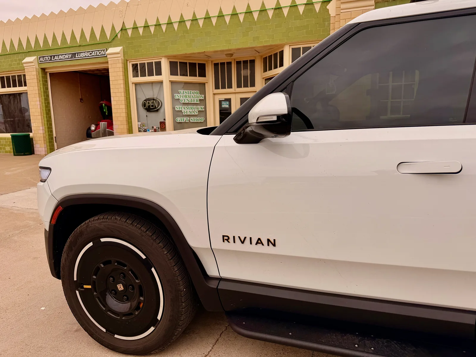 The white Rivian R1T parked at the U-Drop Inn in Shamrock TX, the green-glazed Art Deco tile facade behind with signage reading "AUTO LAUNDRY · LUBRICATION" and "VISITOR INFORMATION CENTER SHAMROCK TEXAS GIFT SHOP", overcast sky